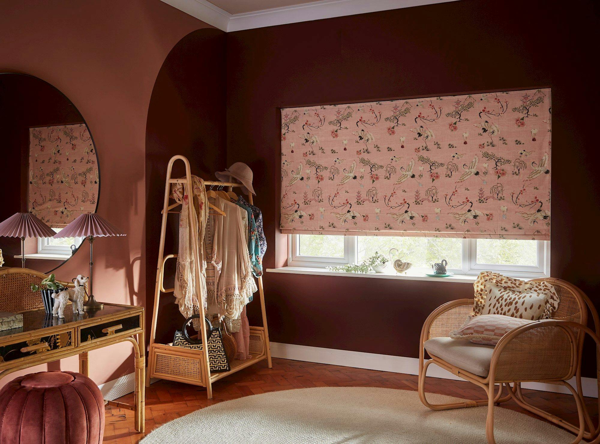 A cozy room features a patterned roller blind over a window, a clothes rack with dresses, a round mirror, a plush ottoman, and a wicker chair with cushions.