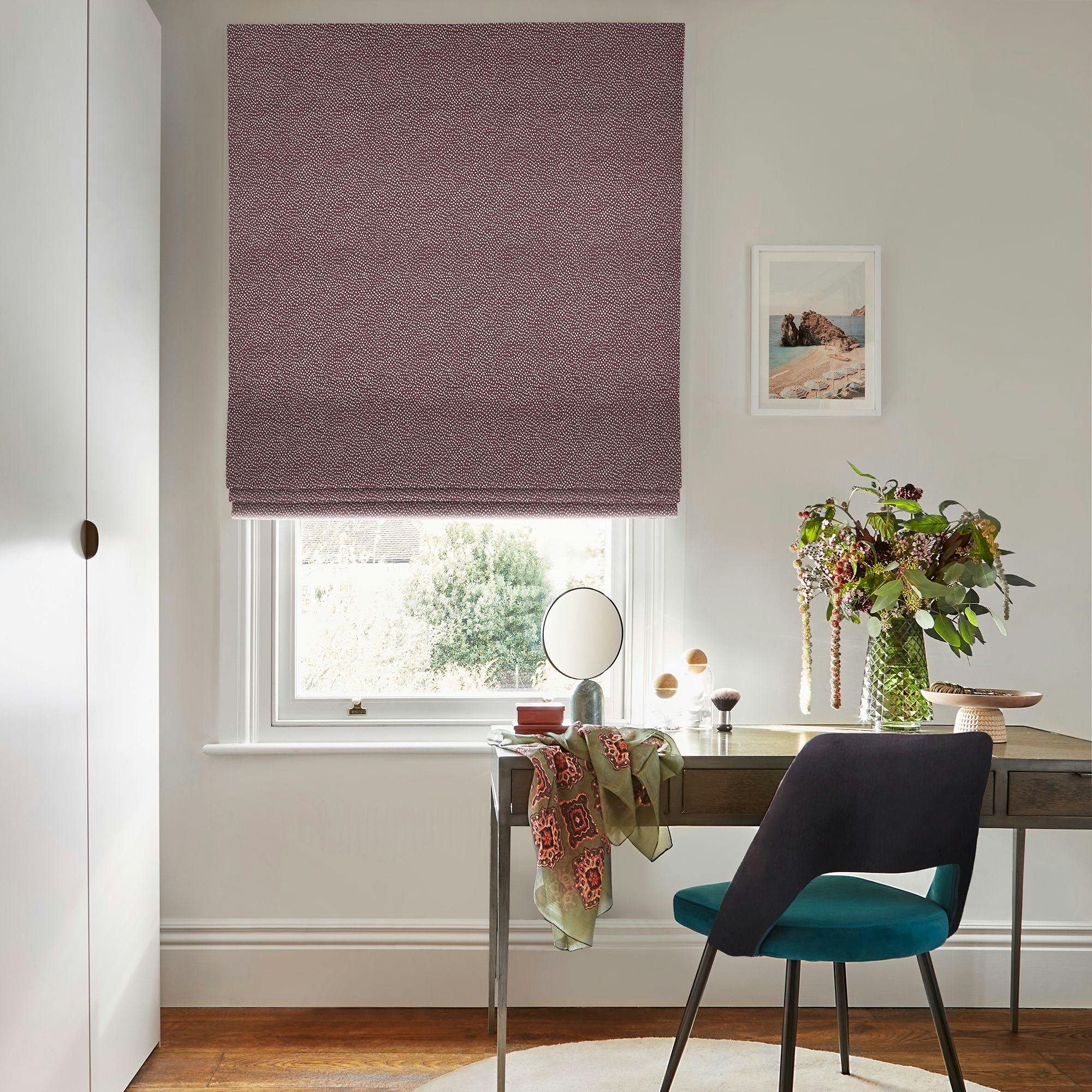 A desk with a decorative scarf and mirror sits in front of a window with a patterned blind. A vase with flowers and framed art enhance the room's ambiance.