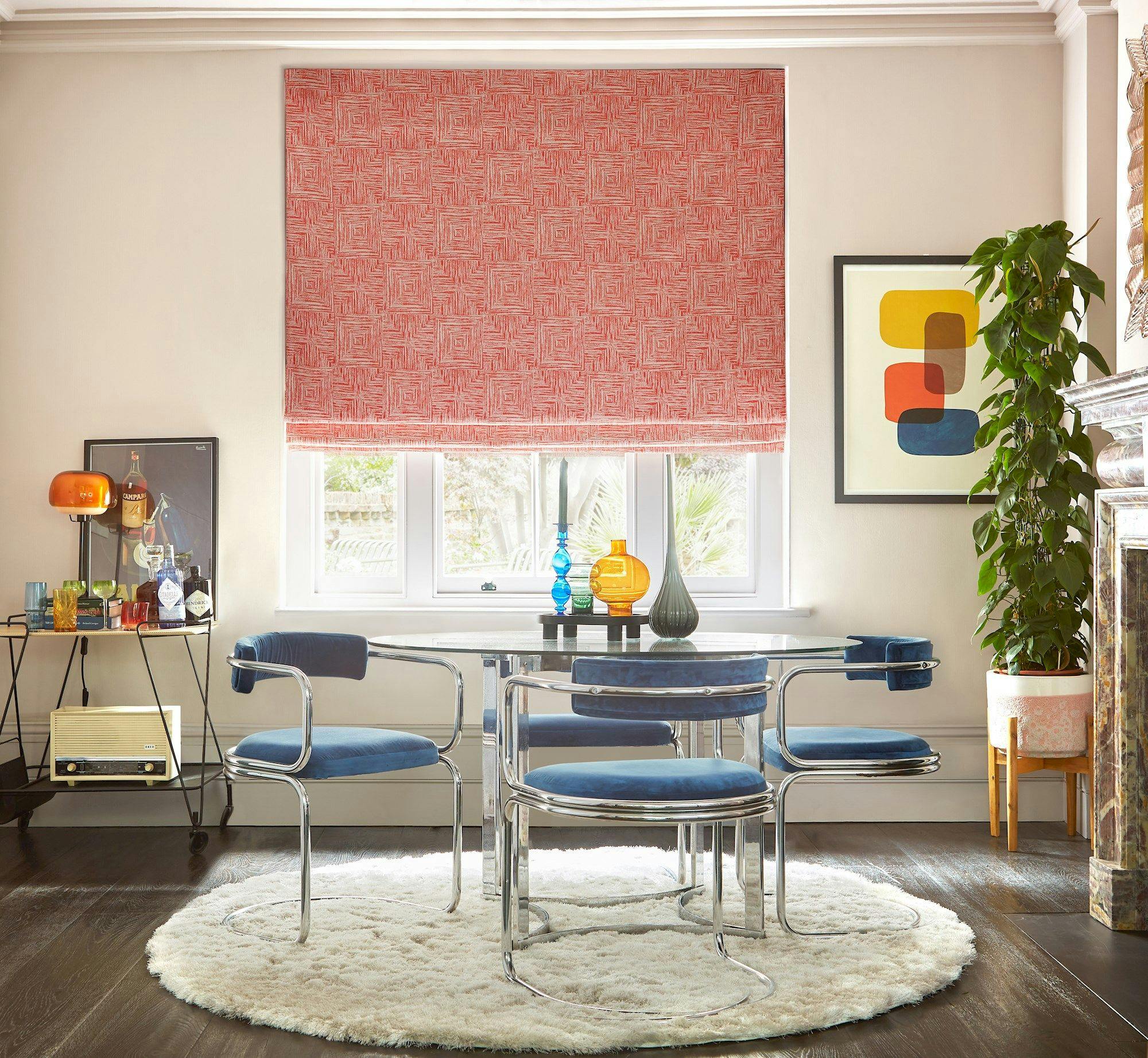 A dining area features a round glass table with chrome and blue chairs, sitting on a circular rug. Nearby, decorative vases and wall art enhance the bright interior space.