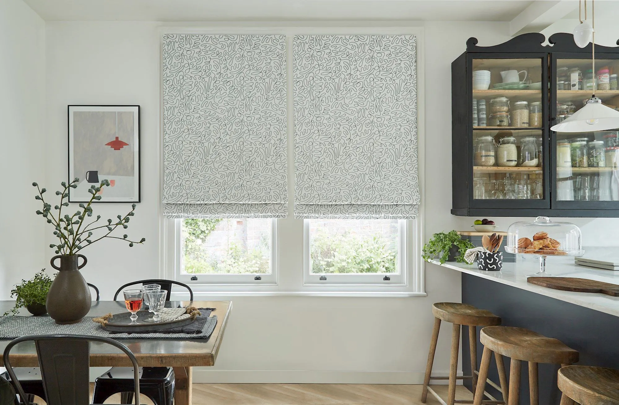 Two patterned Roman blinds hang over windows, partially covering outdoor greenery. A kitchen features a wooden table with a vase, framed art, and neatly arranged chairs. Jars sit in a nearby cabinet.