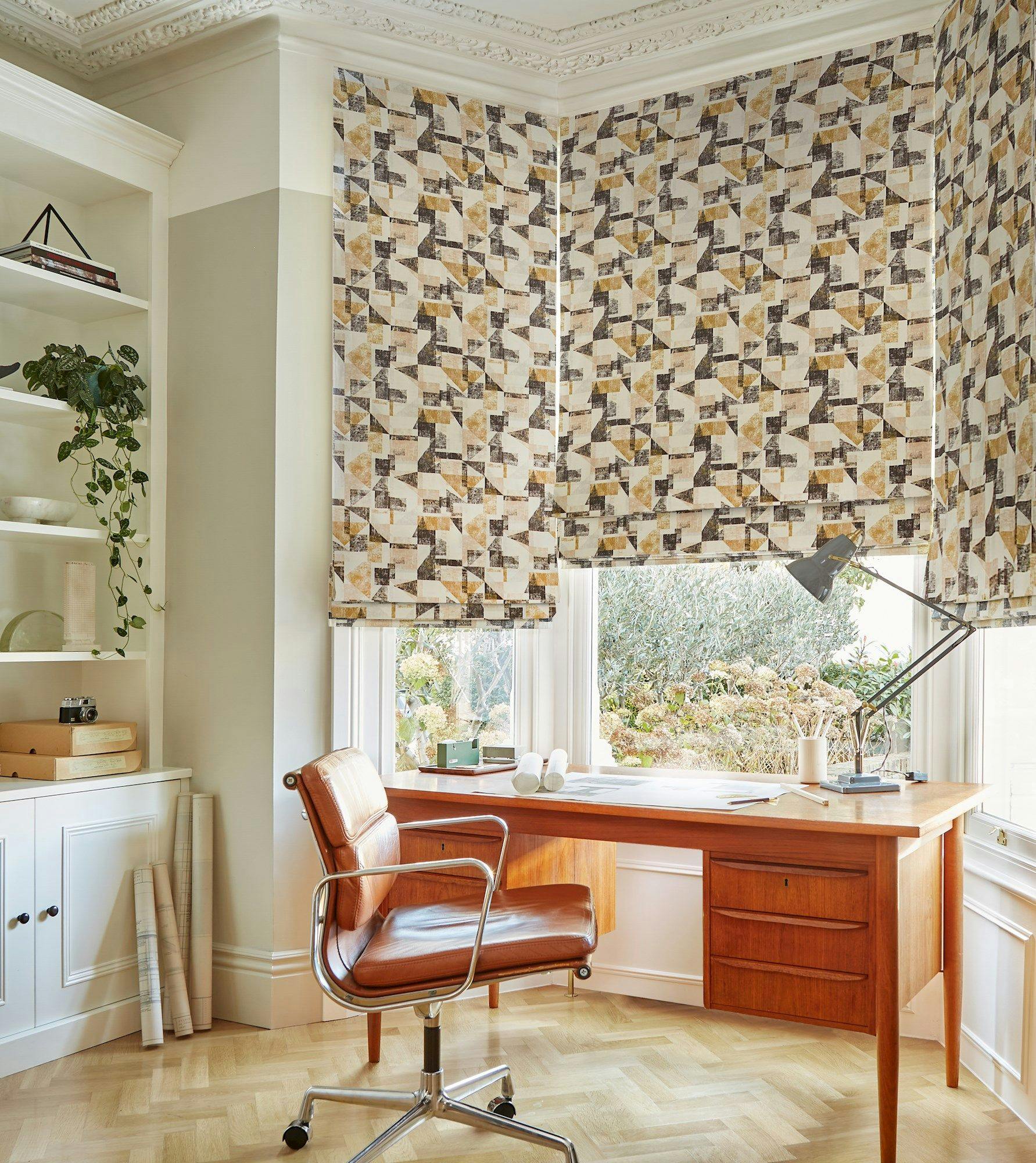 A wooden desk holds a lamp and papers, positioned near patterned window shades. A brown swivel chair faces the desk. Shelving with books and plants decorates the room's corner.