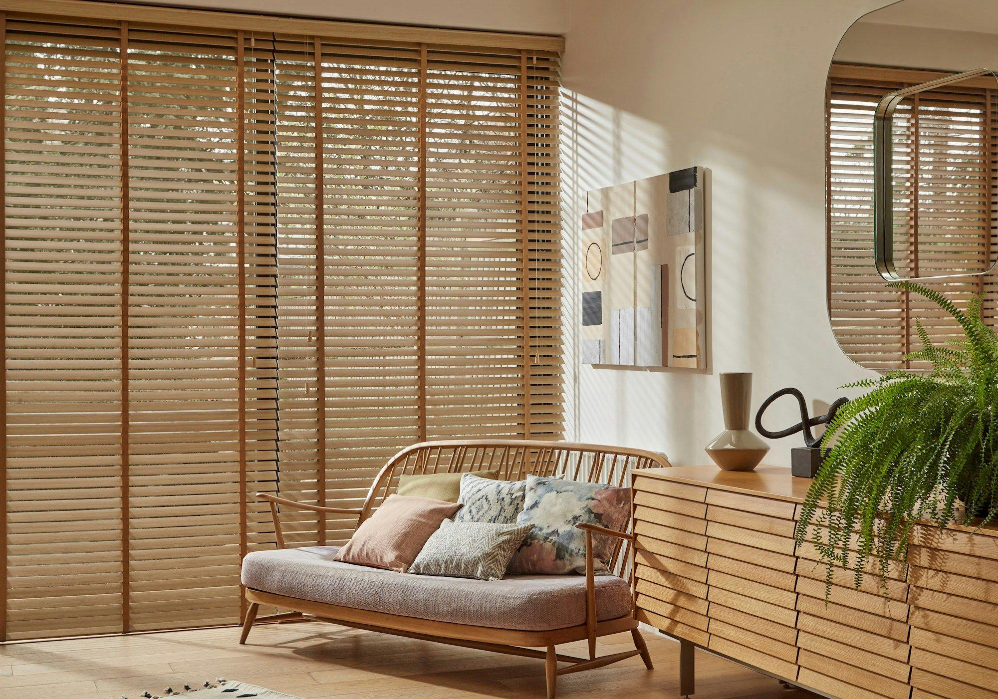 A wooden sofa with colorful cushions sits in a bright room. Sunlight filters through slatted blinds. Wooden furniture and abstract artwork decorate the space, alongside a green plant.