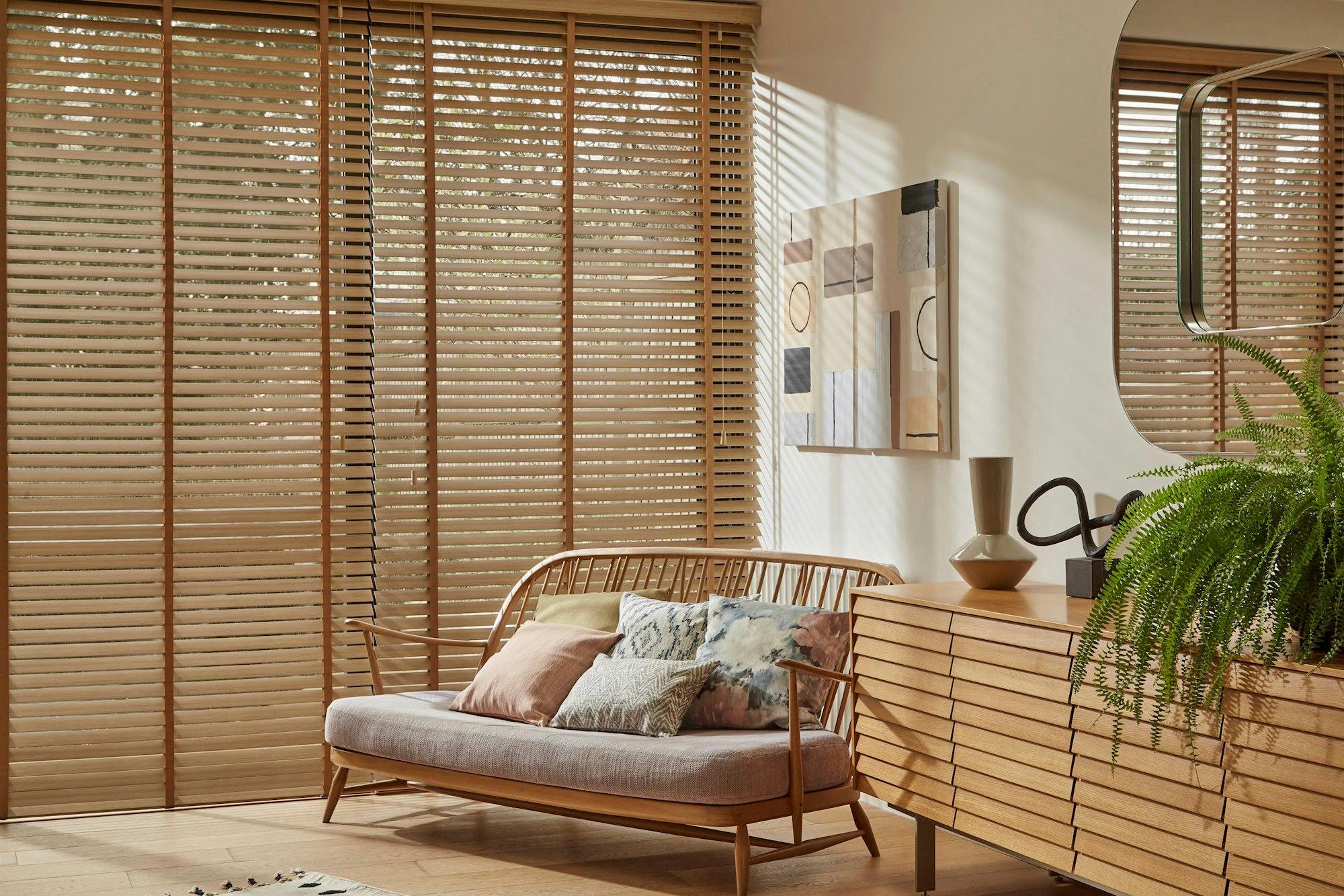 A wooden sofa with colorful cushions sits in a bright room. Sunlight filters through slatted blinds. Wooden furniture and abstract artwork decorate the space, alongside a green plant.