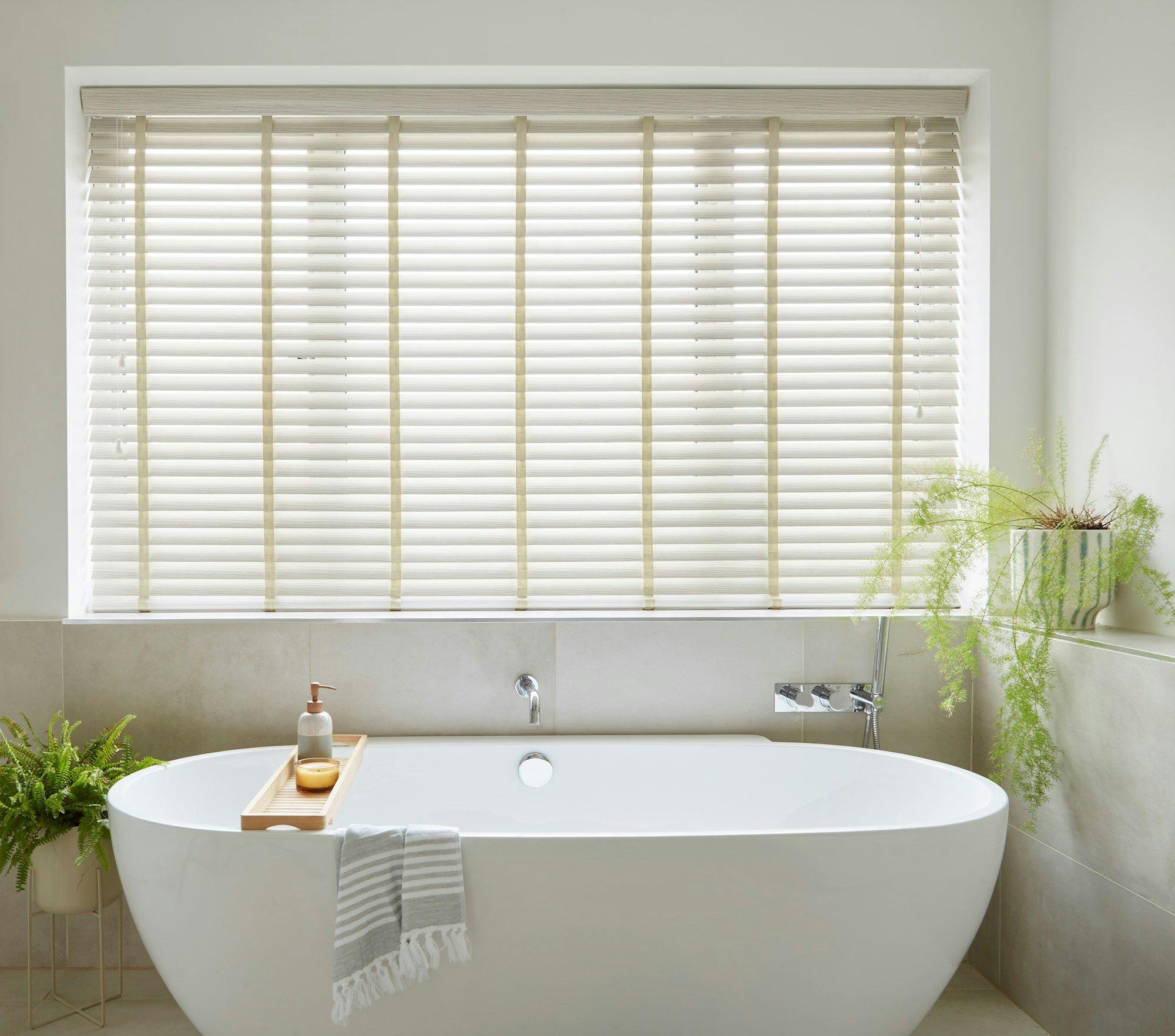 Bathtub stands filled in a modern bathroom, featuring a wooden tray with soap and candle. White blinds cover a large window, surrounded by green plants on the floor and ledge.