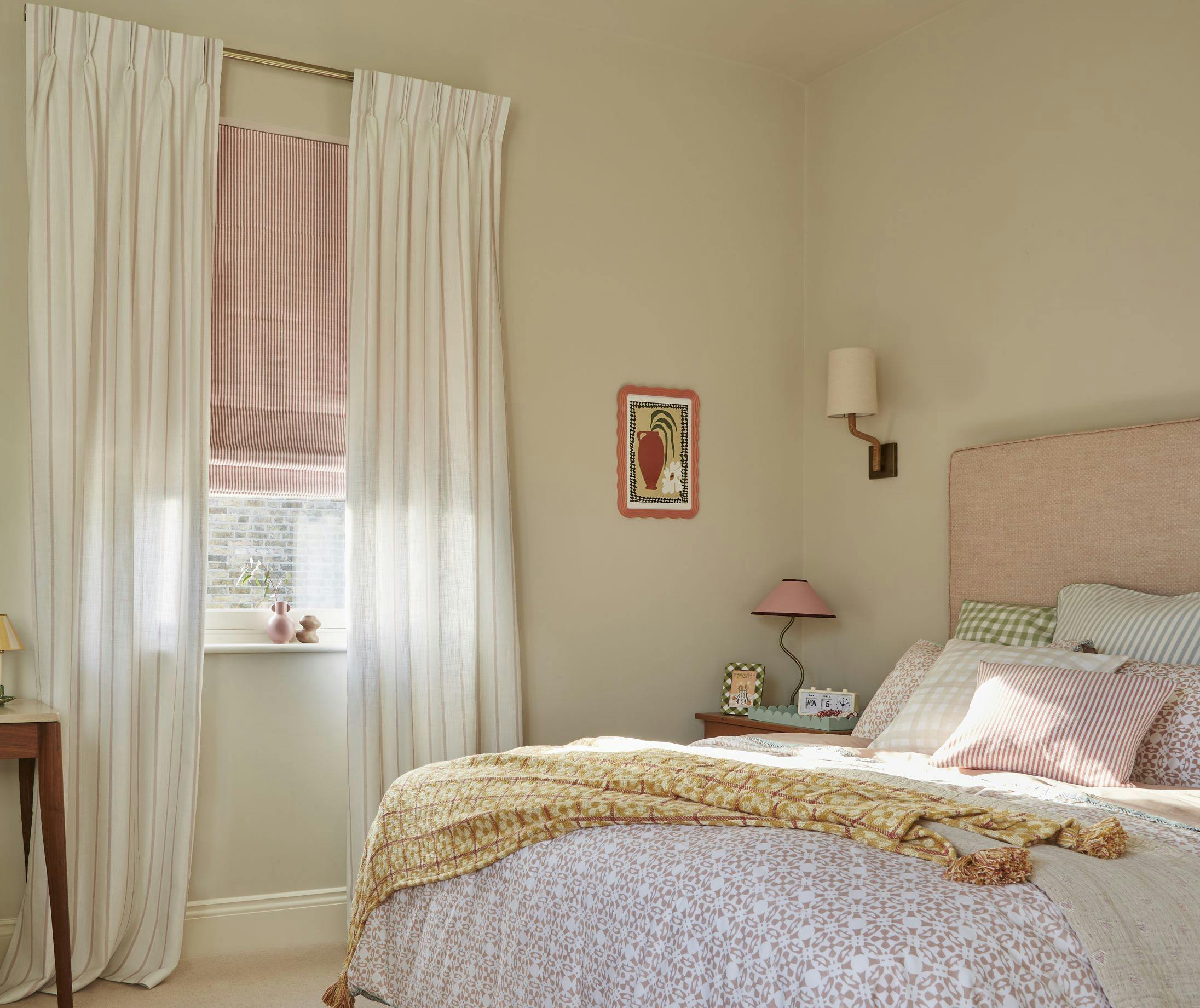 A cozy bedroom with a bed covered in patterned bedding, a pink headboard, and assorted pillows. A window with soft curtains and a pink blind lets in light. A side table holds framed photos and a lamp.