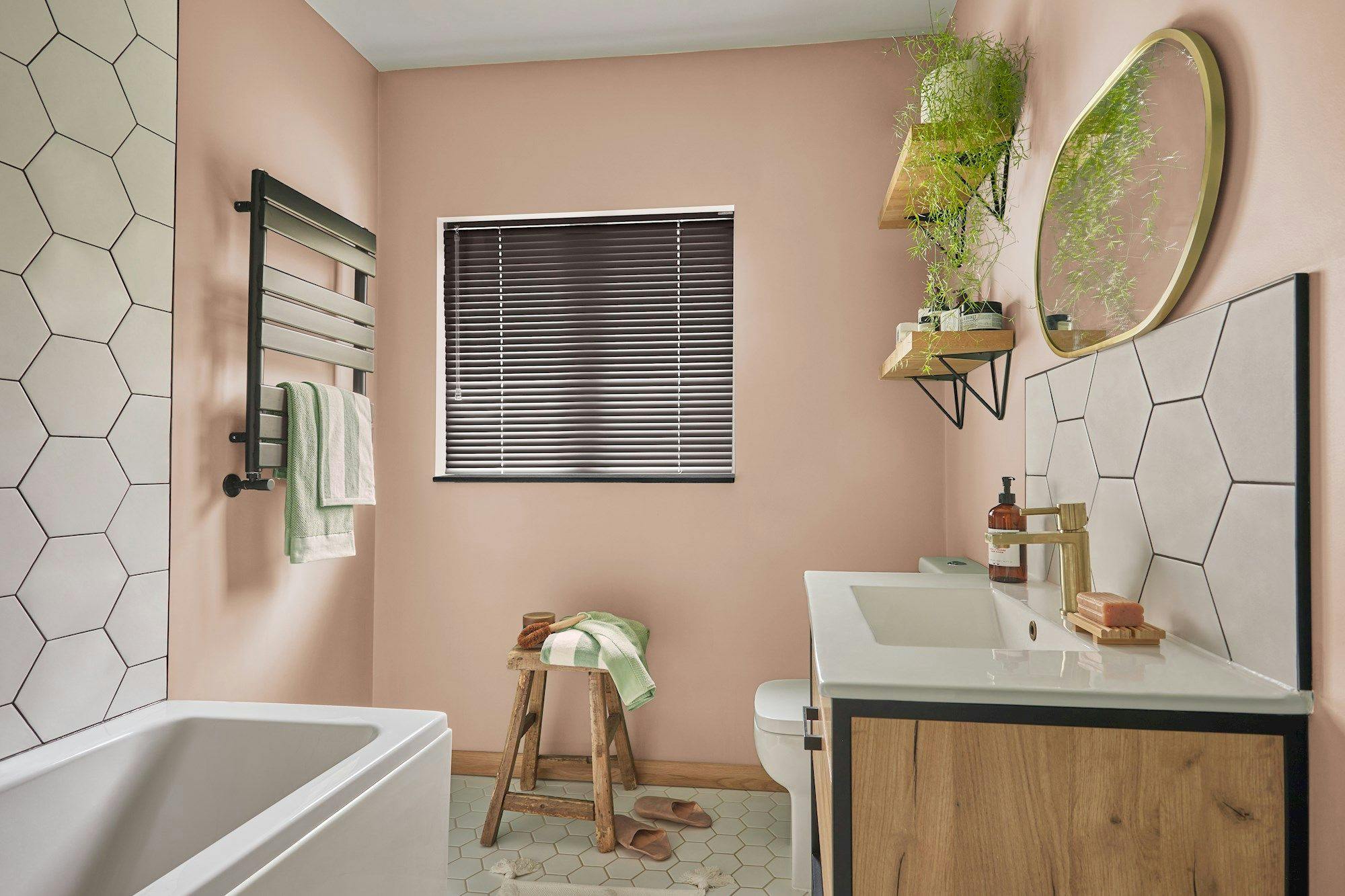 Bathroom with a white bathtub, wooden stool holding towels, and hanging plant above a wooden vanity. Hexagonal tiles and a window with blinds create a modern, cozy atmosphere.