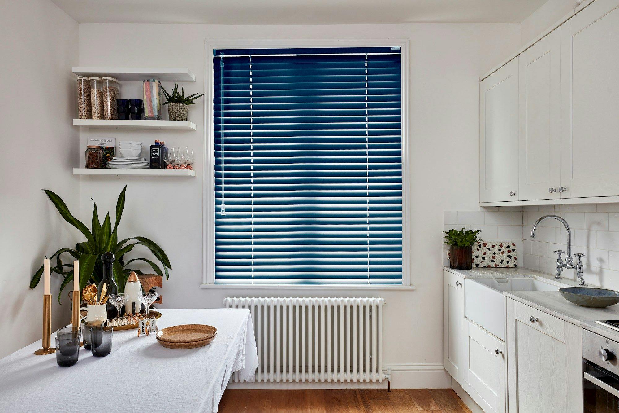 Table set with glassware and candles next to potted plant. Blue blinds cover window above radiator. Shelves hold jars and decor. White cabinets and sink occupy the right.