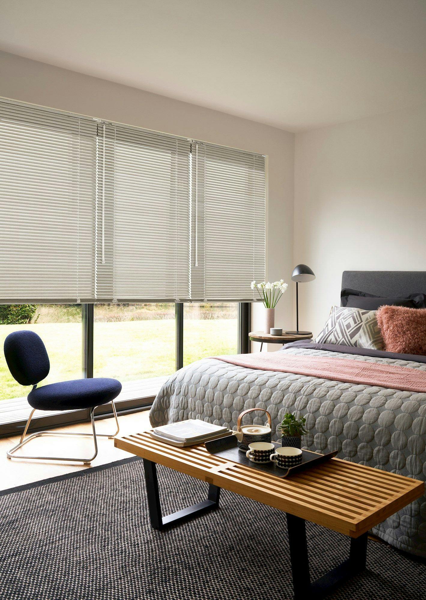 A modern bedroom contains a bed with textured bedding, a wooden bench with tea set, a blue chair, and a side table with flowers, all under large windows with blinds.