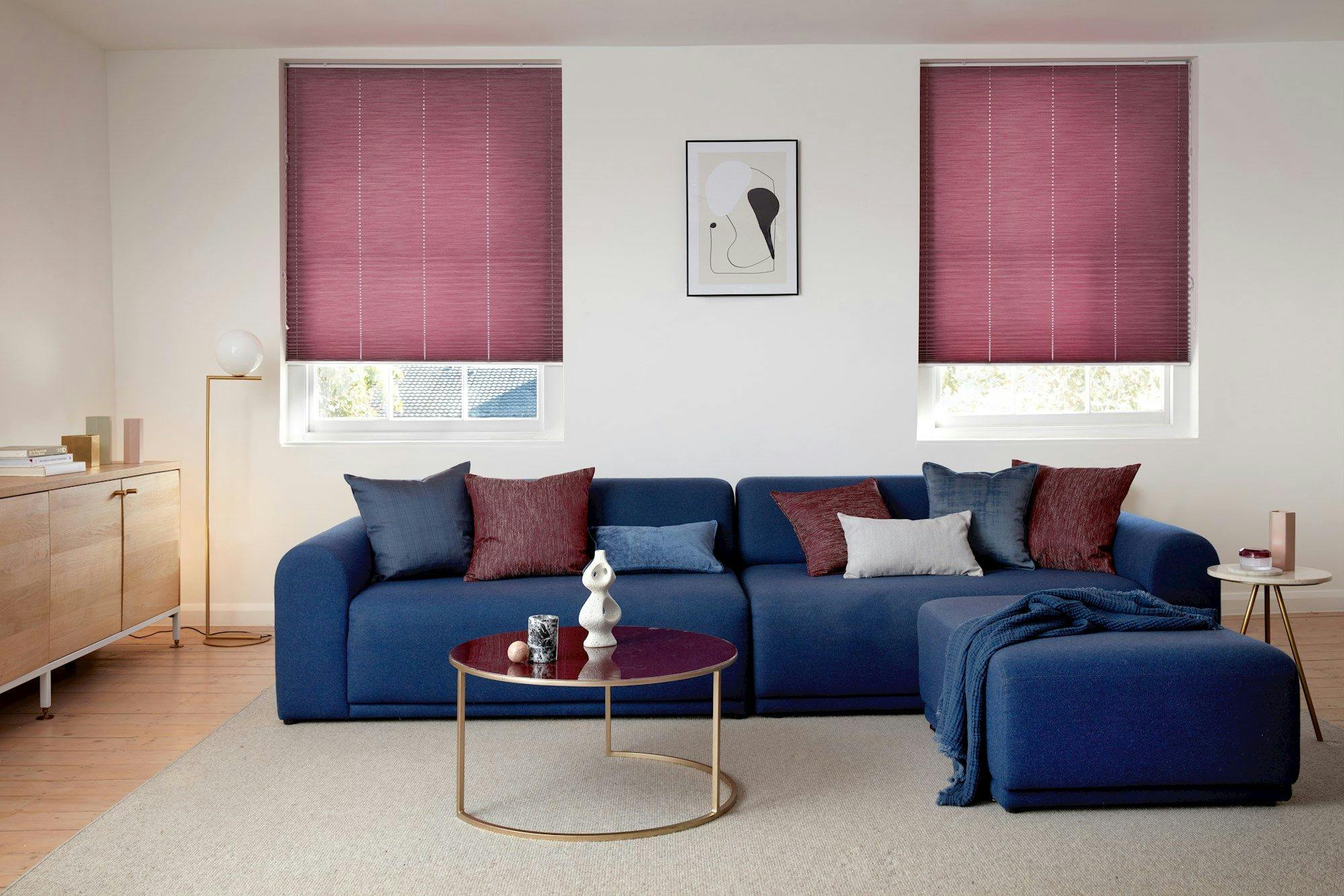 A blue sofa with assorted pillows rests against a white wall. Two windows with red blinds flank a framed abstract print. Wooden flooring, a light, and side tables complete the room.