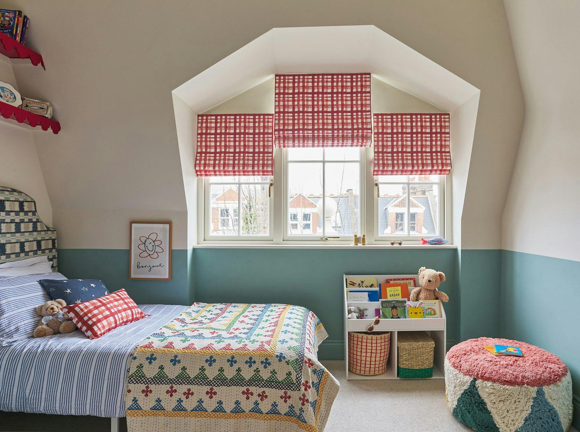 A cozy bedroom features a bed with striped bedding and a colorful quilt, a teddy bear sitting on it. Books and toys are on shelves under a window with red-checkered blinds. A picture with a flower and the word �bonjour� is on the wall.