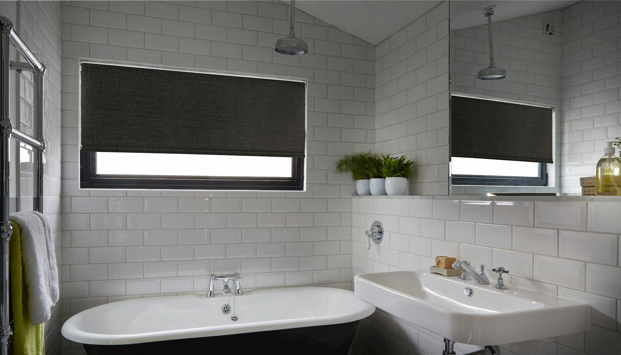 Bathtub sits beneath a window with a dark roller blind, surrounded by white subway tiles. Nearby, a sink and potted plants add decor, while a towel hangs on a heated rail.