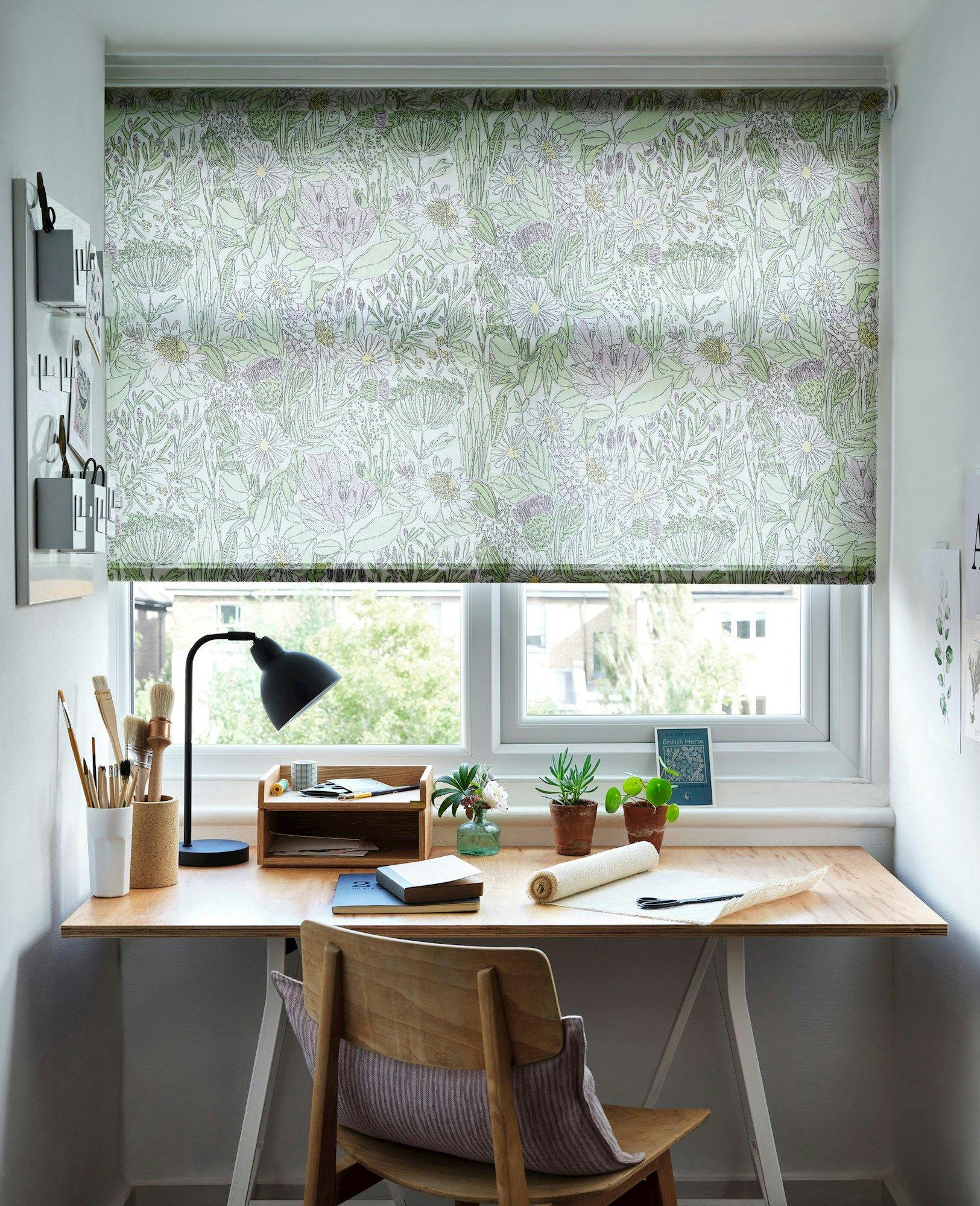 A wooden desk with art supplies and a lamp faces a window with floral-patterned blinds. Chairs and plants add to the cozy aesthetic, emphasizing a creative workspace.