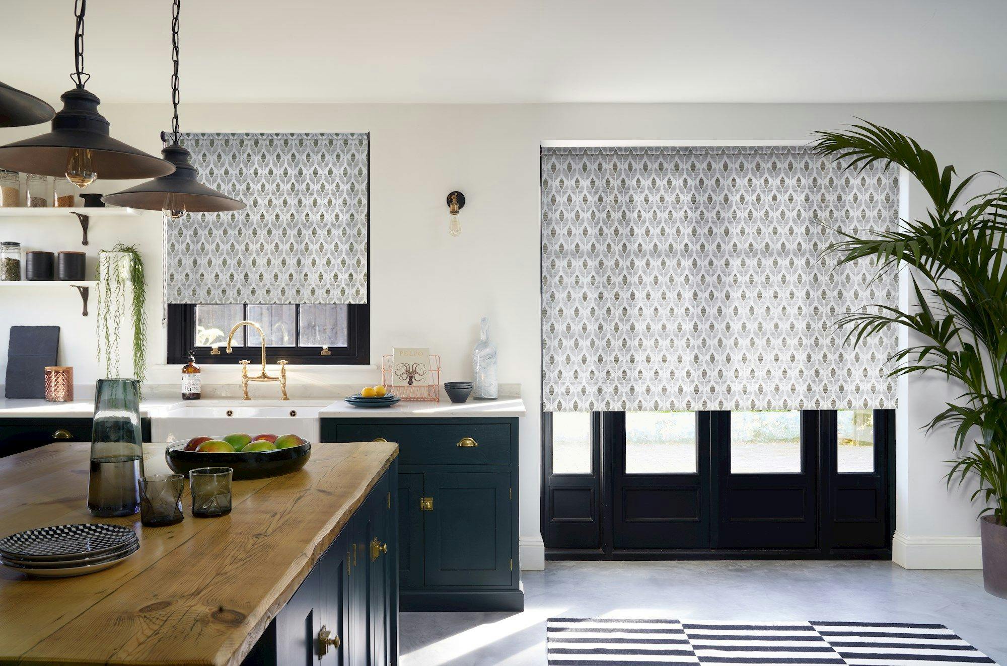 Blinds cover windows above a kitchen sink and double doors. A wooden counter holds fruit, plates, and glasses. Shelves display jars; a potted plant stands nearby.