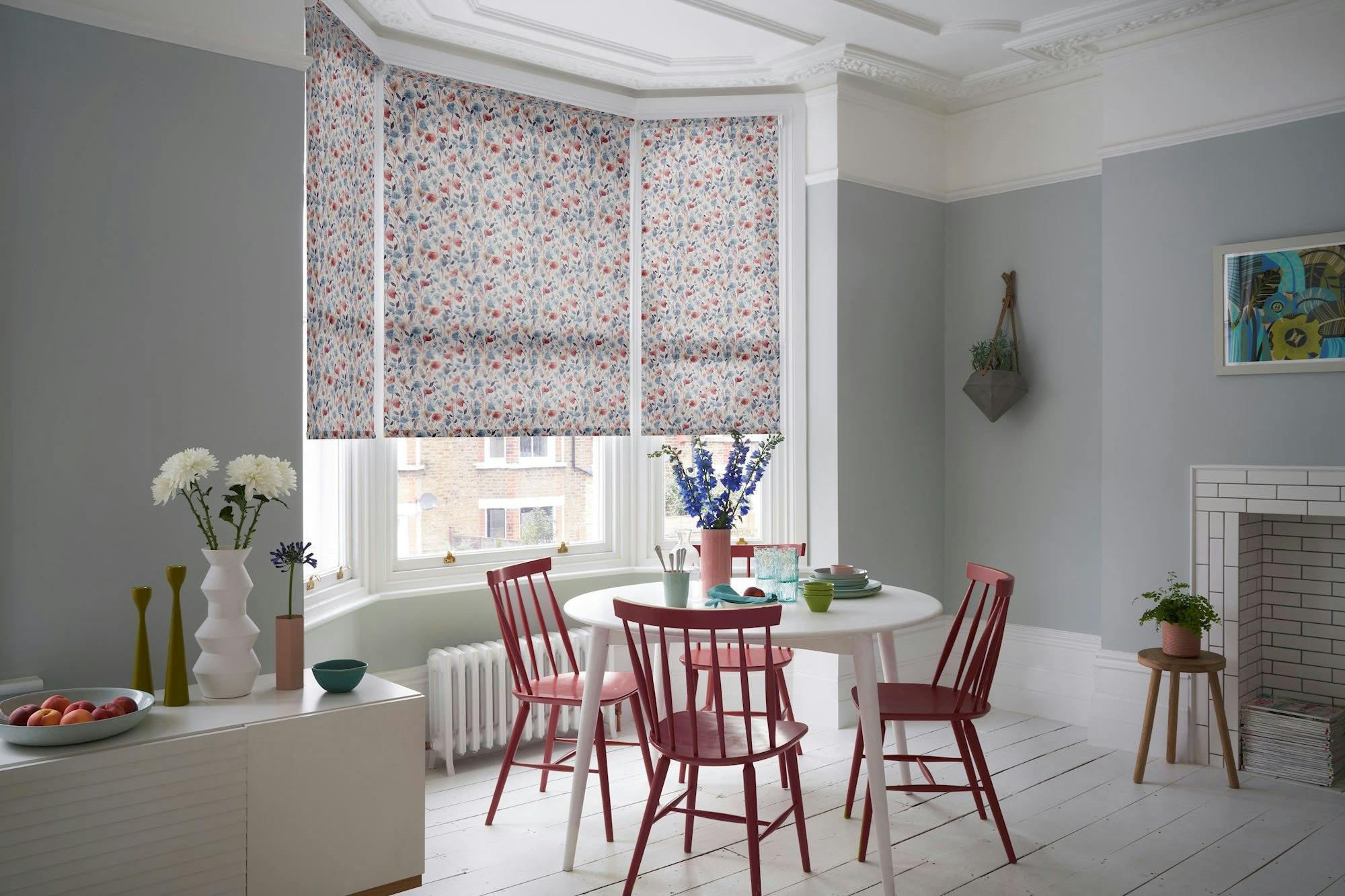 A dining table with red chairs sits in a room with floral roller blinds and pastel walls. Decorative vases and plants add accents, while a fireplace and modern art complete the setting.