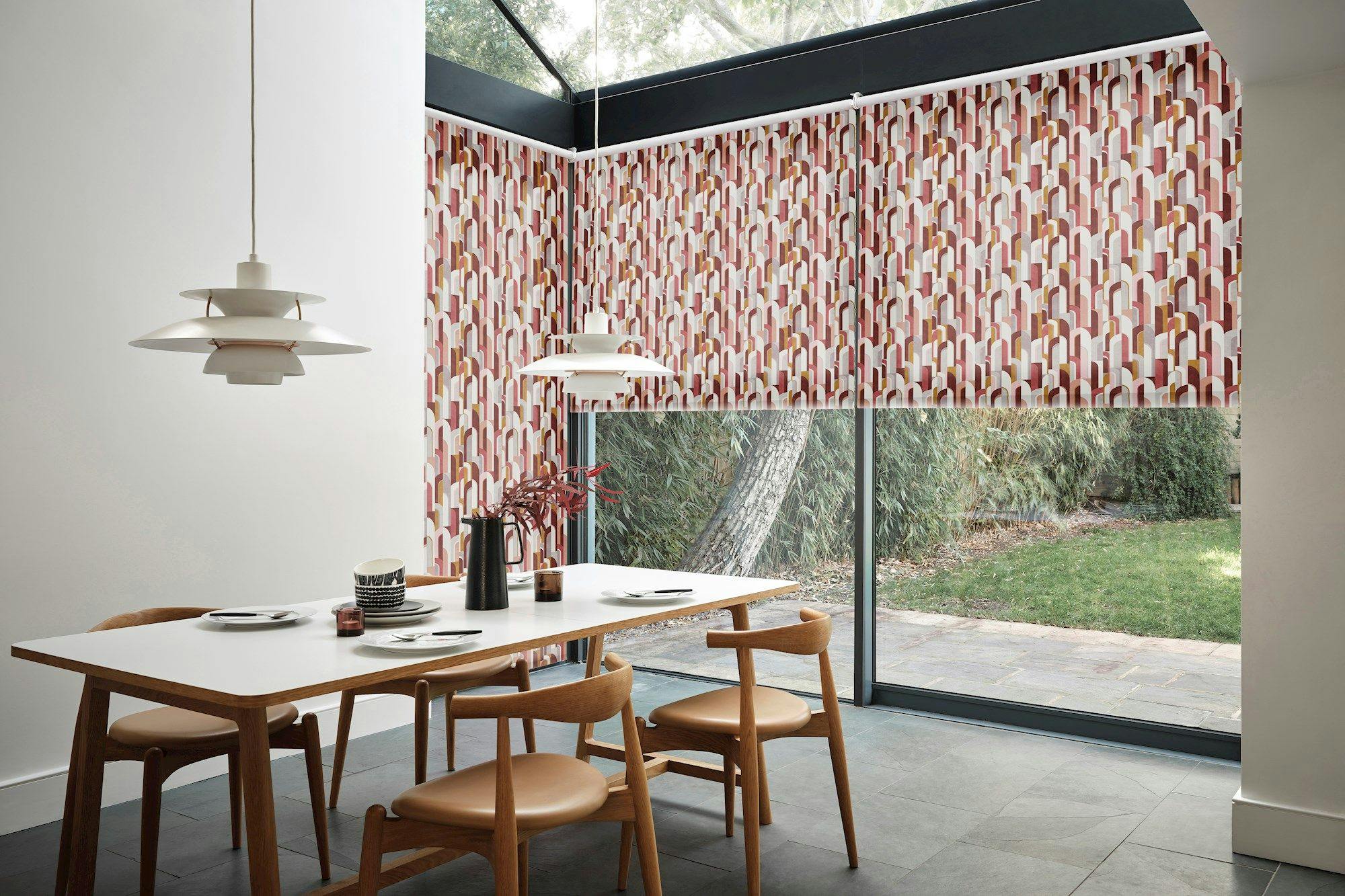 A modern dining table with wooden chairs holds plates and a jug. Overhead, pendant lamps hang. Roller blinds with red and white patterns cover glass walls, revealing a garden outside.