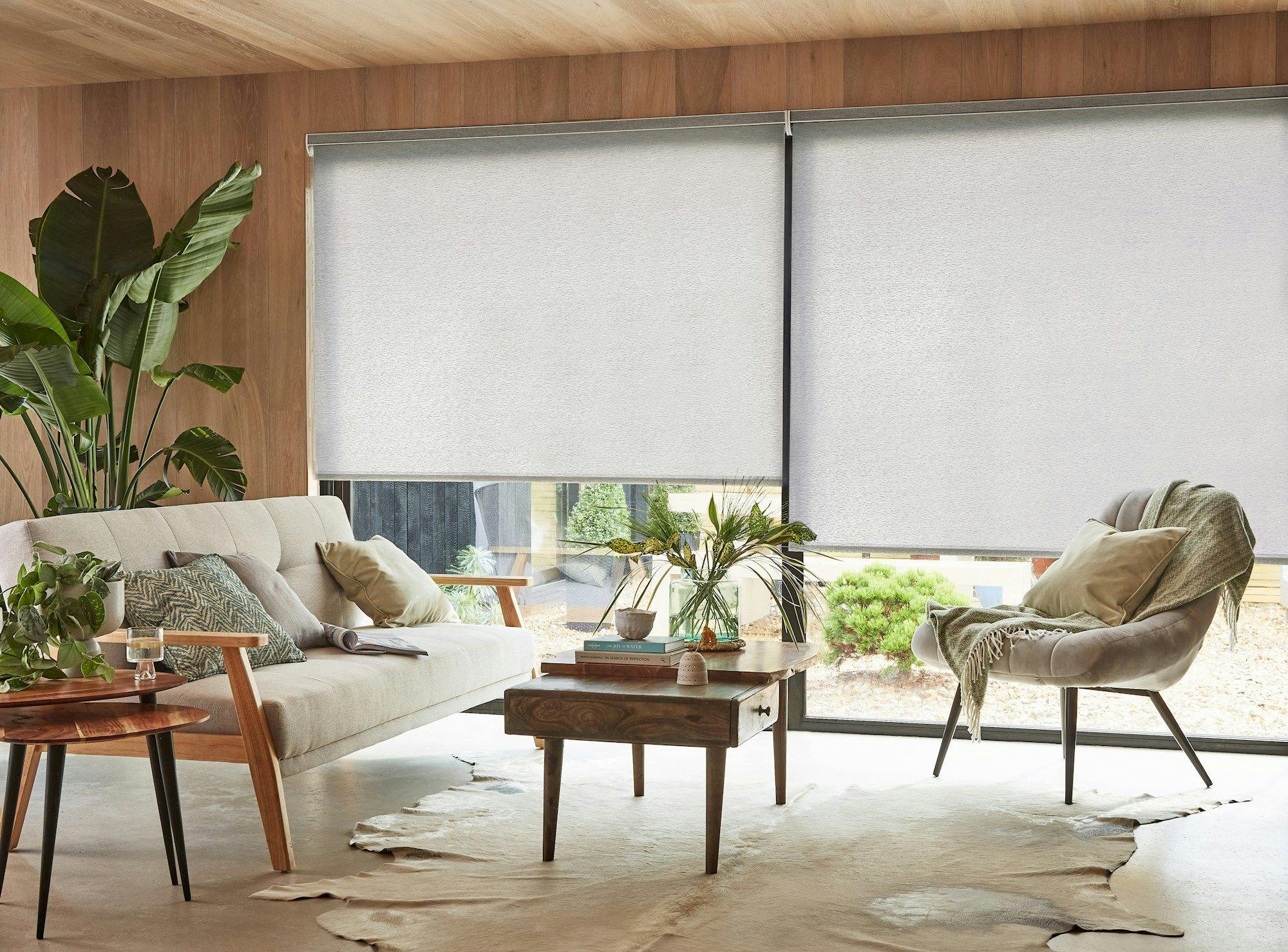 Sofa and chair adorned with cushions and blanket, placed around a wooden coffee table; indoor plants are beside a large window with closed blinds in a cozy living room.
