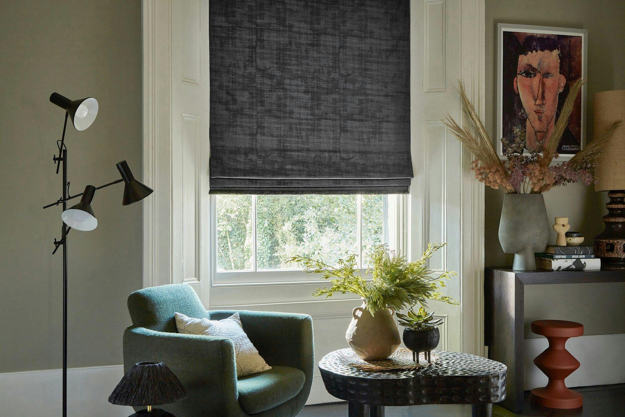 A teal armchair with a cushion sits near a window with closed dark blinds. Nearby, a table holds vases with plants. A framed portrait hangs above decorative items on a console.