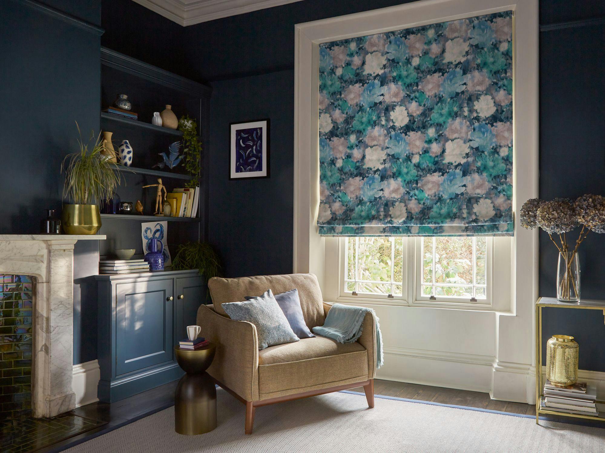 A beige armchair with cushions sits by a side table with a cup in a cozy room. A floral patterned blind covers the window, and shelves with books and decor accent the dark blue walls.