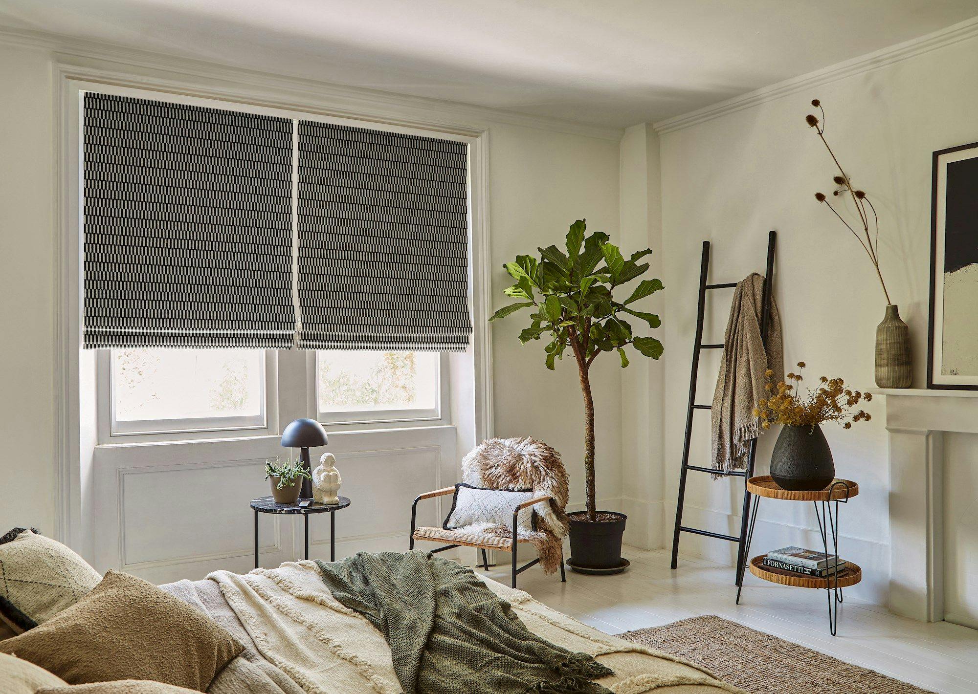 Room with a bed covered in neutral linens. Black Roman shades cover windows. Plant, chair, and ladder decor in corner. Lamp on side table near window and bed.