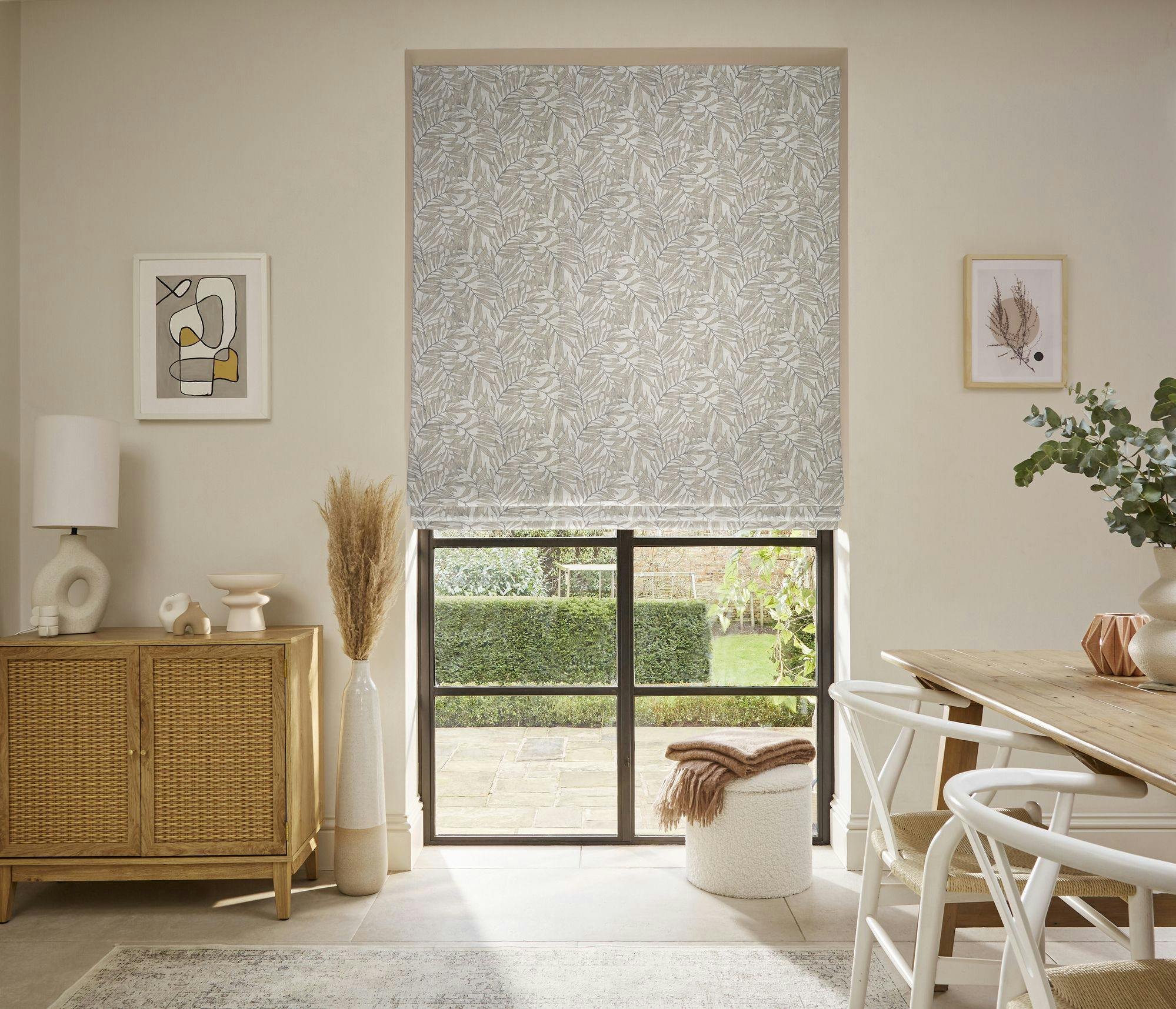 A light, airy living room showcases a large window with leafy patterned blinds. A wooden sideboard holds decor, while a dining table with white chairs and green plants adds coziness.