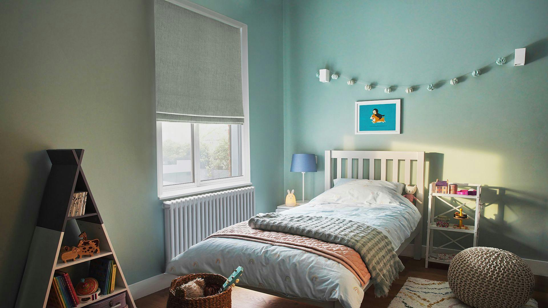 A neatly made bed with a blue lamp on the bedside table sits in a pastel-colored room. String lights hang on the wall, with a toy shelf and pouf nearby. Text not present.