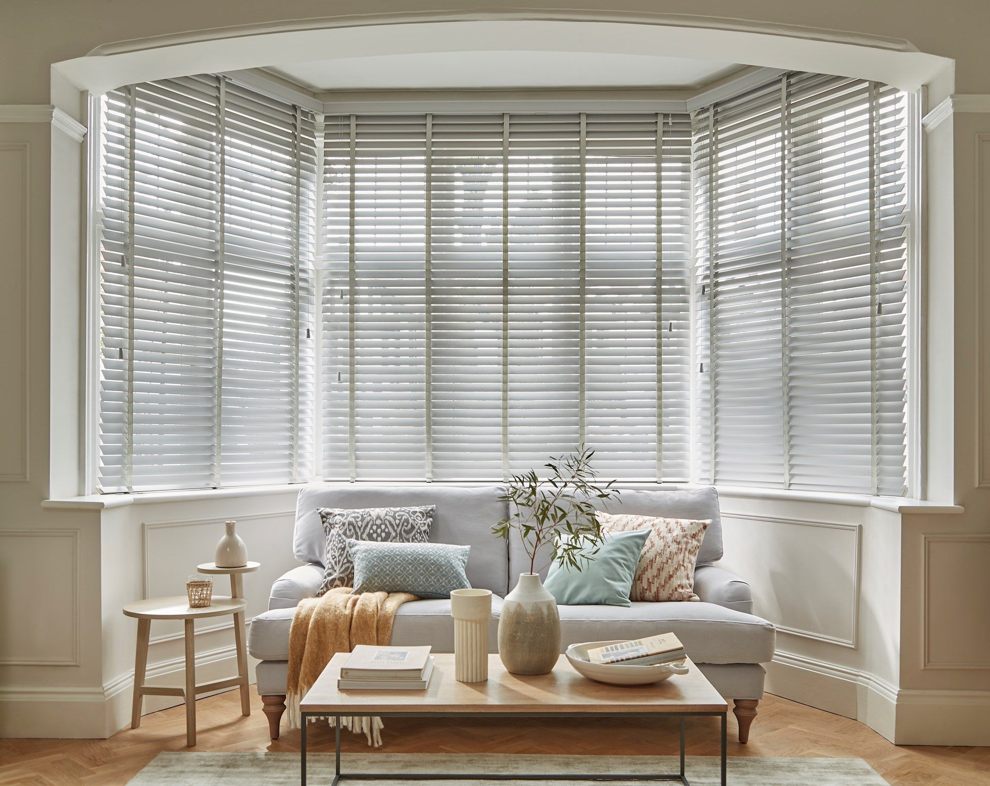 A cozy living room features a sofa adorned with patterned cushions and a throw blanket, surrounded by light-filtering white blinds on bay windows, with wooden side tables and decorative items.