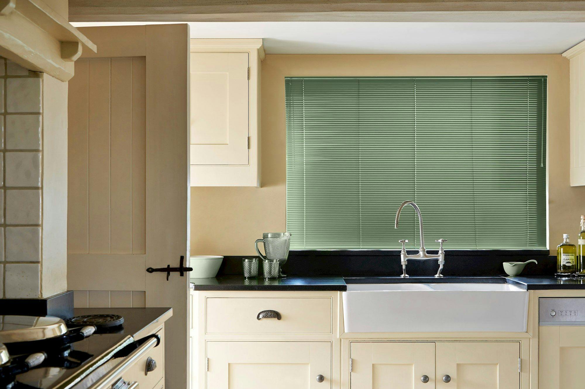 A farmhouse-style kitchen features a ceramic sink beneath green blinds, surrounded by cream cabinets. A partially open wooden door and countertop accessories complete the cozy setting.