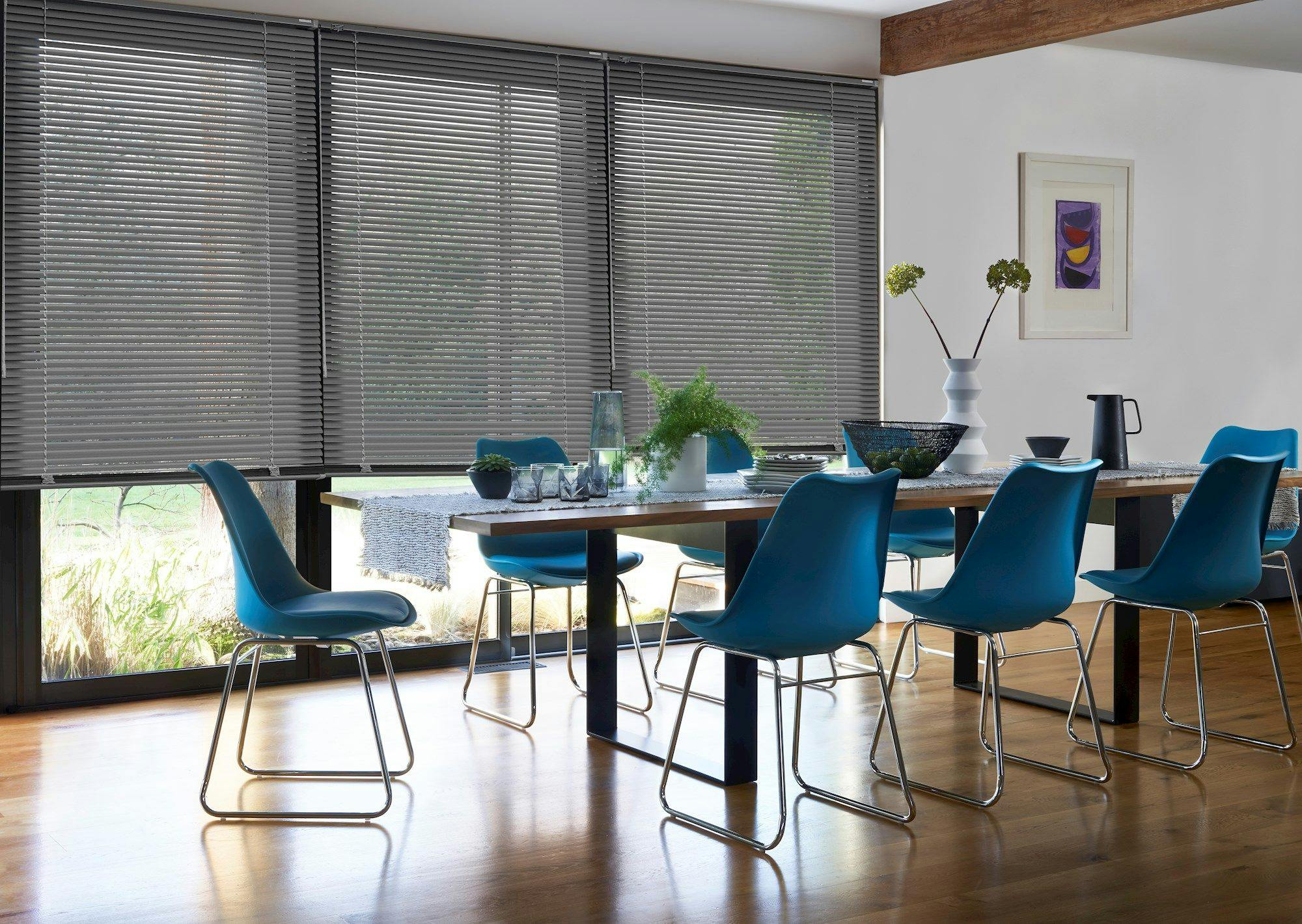 A long dining table with blue chairs is set with dishes and plants in a room featuring wooden floors, modern decor, and large windows with closed blinds.