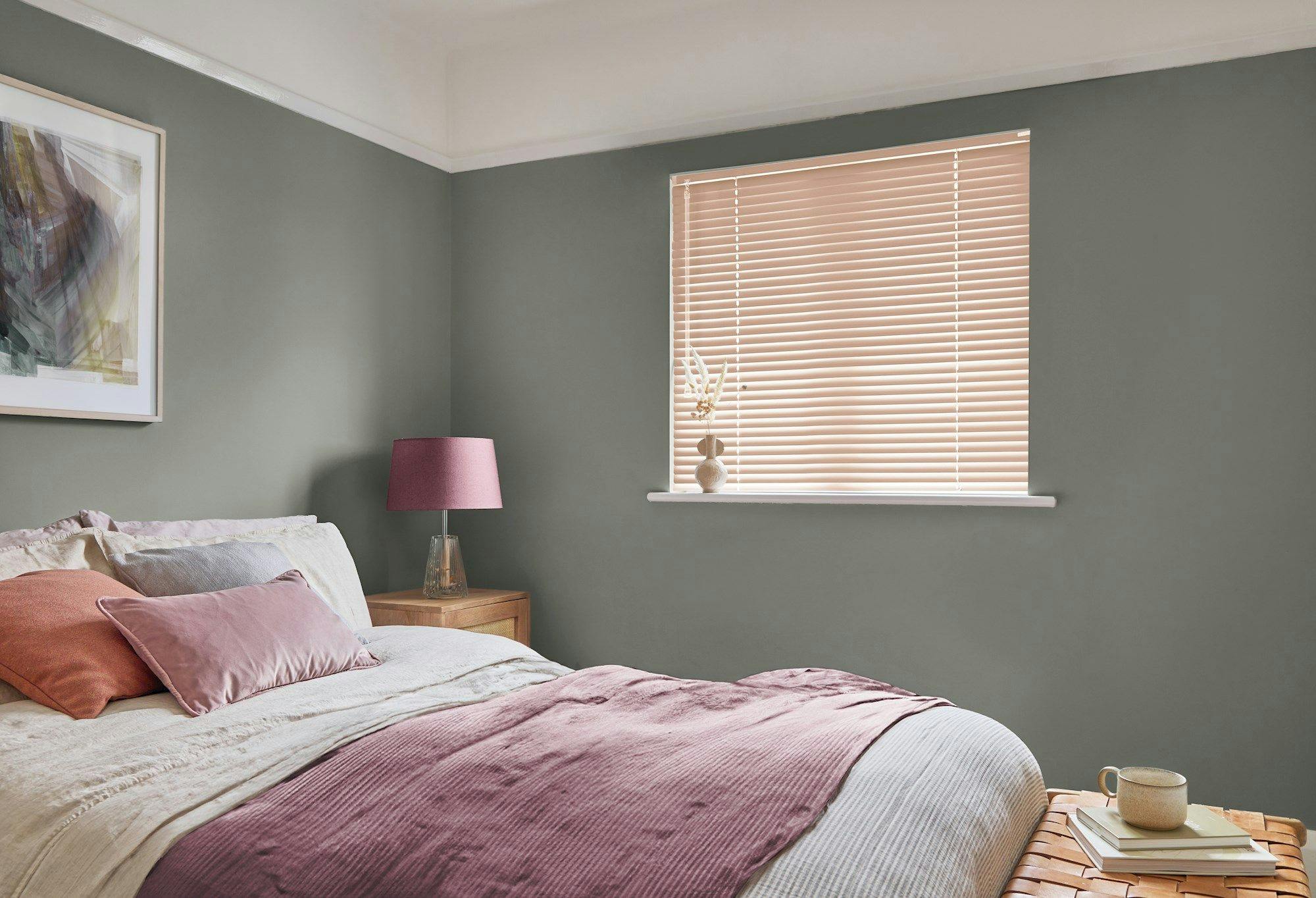 Bedroom with a neatly made bed featuring pink and beige bedding, adjacent to a nightstand with a pink lamp. Soft green walls, a window with blinds, and a framed abstract artwork.