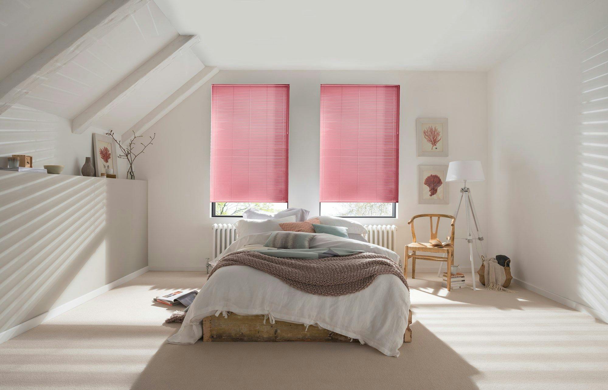 A cozy bedroom features a bed with white and brown bedding, under two windows with pink blinds, surrounded by light walls, decorative plants, and framed art.