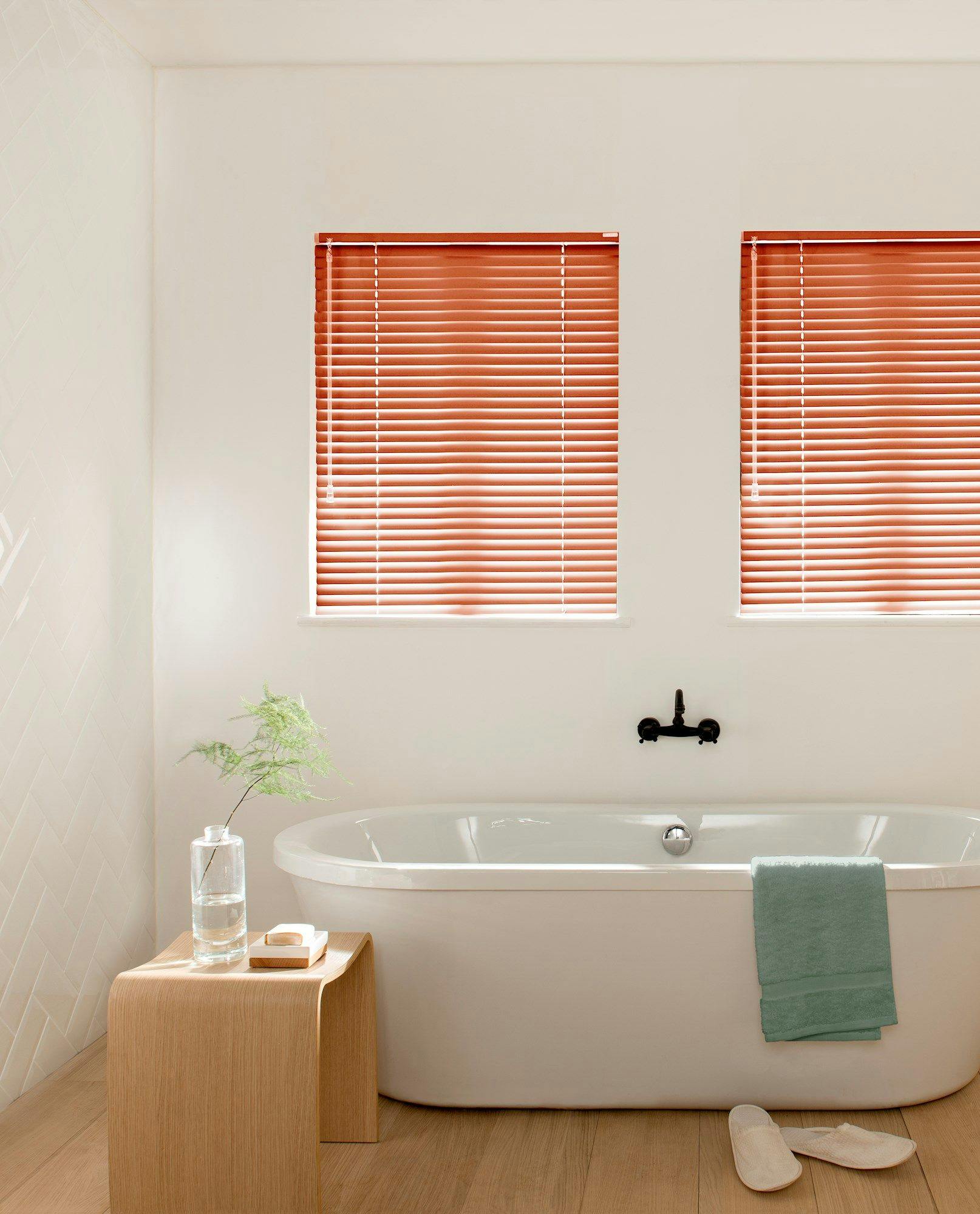 A white bathtub sits under closed red blinds, flanked by a wooden stool with a vase and soap. Soft wood flooring complements green towels, with white slippers nearby.