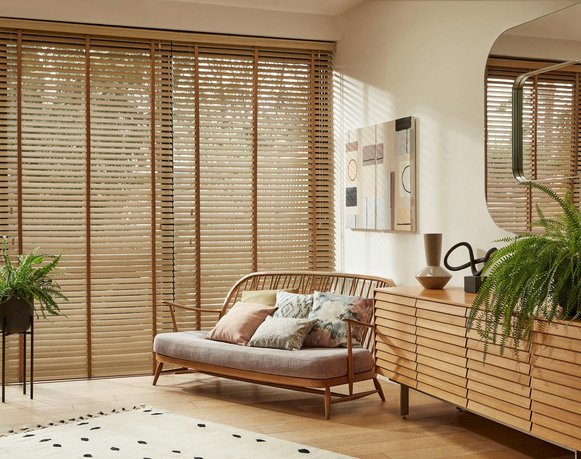 Wooden sofa with cushions sits in front of large windows with wooden blinds. Nearby, a wooden sideboard holds decorative items, surrounded by potted ferns and abstract artwork on the wall.