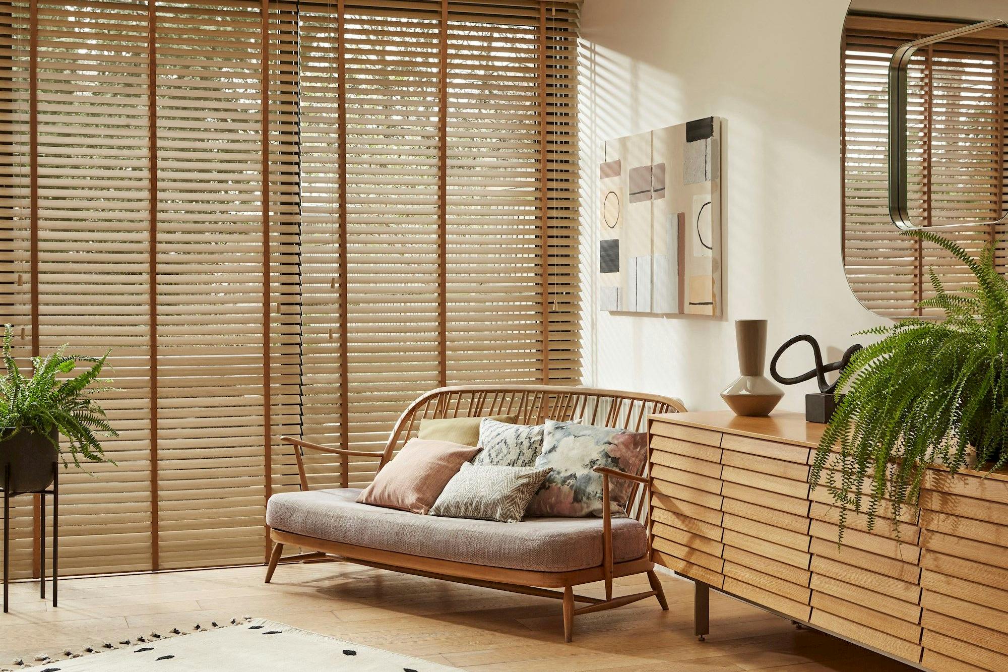 Wooden sofa with cushions sits in front of large windows with wooden blinds. Nearby, a wooden sideboard holds decorative items, surrounded by potted ferns and abstract artwork on the wall.