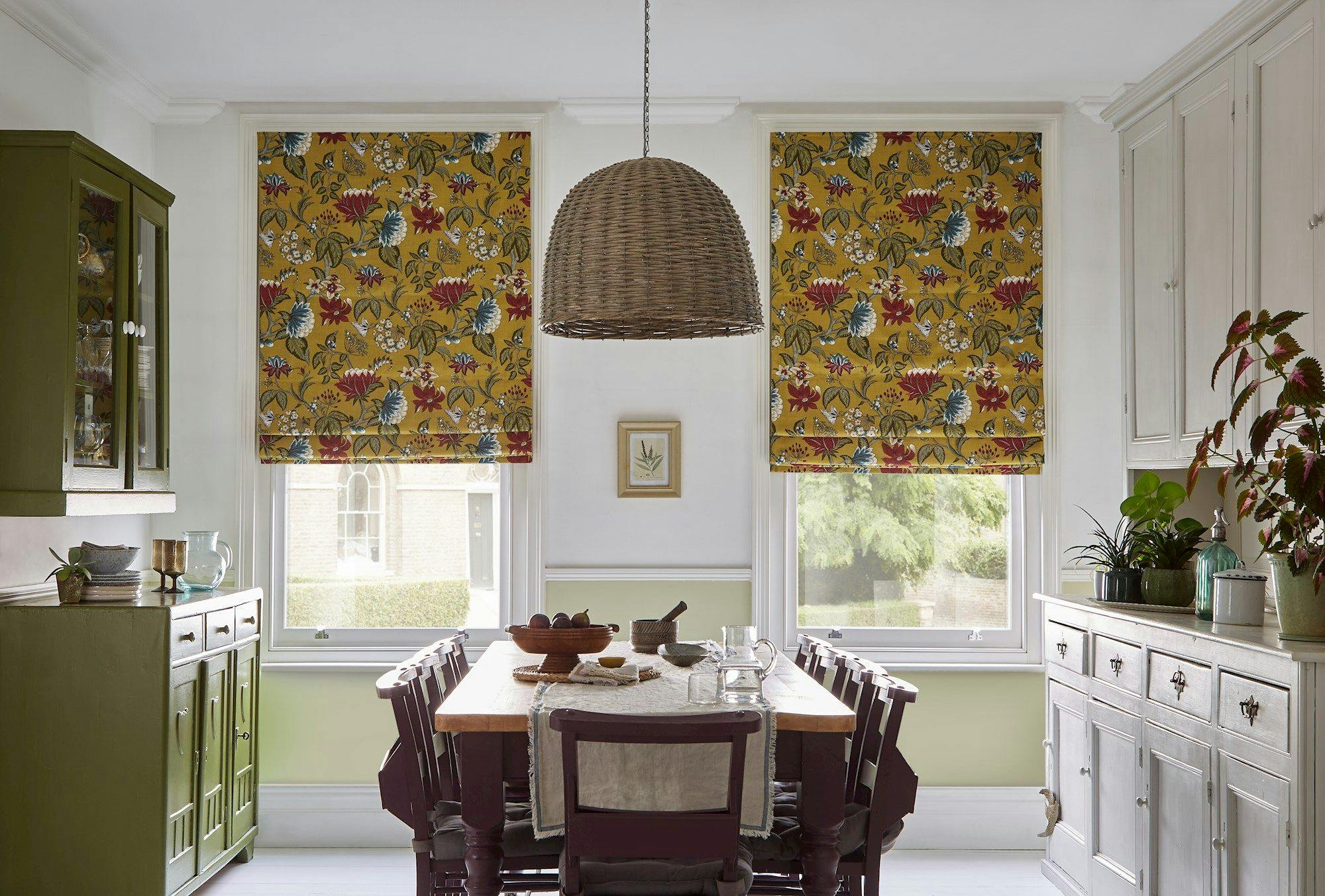 Dining table set with dishes and wooden bowls, beneath a woven pendant light, surrounded by cabinets and floral Roman shades, in a bright, plant-decorated kitchen.