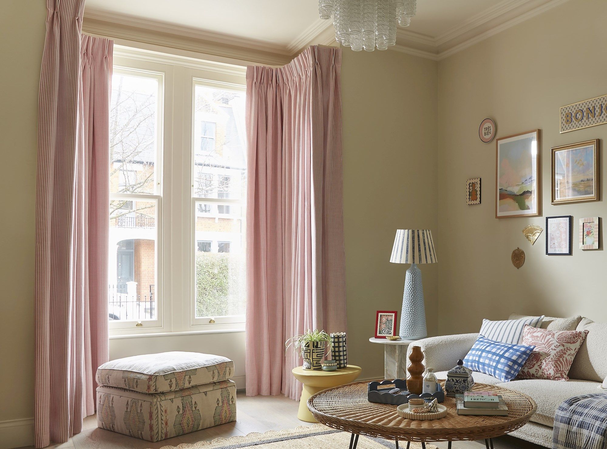 A cozy living room features pink curtains framing tall windows. A patterned ottoman and wicker table sit near a sofa with cushions. Various framed artworks adorn the light-colored walls.