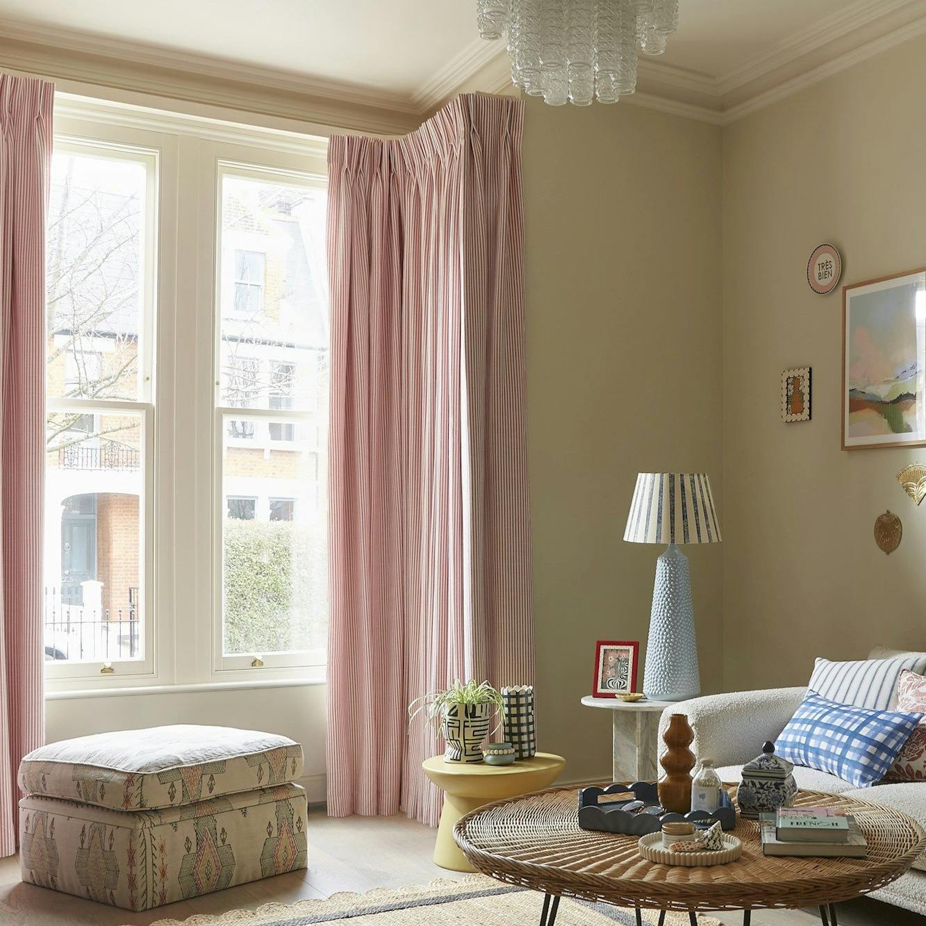 A cozy living room features pink curtains framing tall windows. A patterned ottoman and wicker table sit near a sofa with cushions. Various framed artworks adorn the light-colored walls.