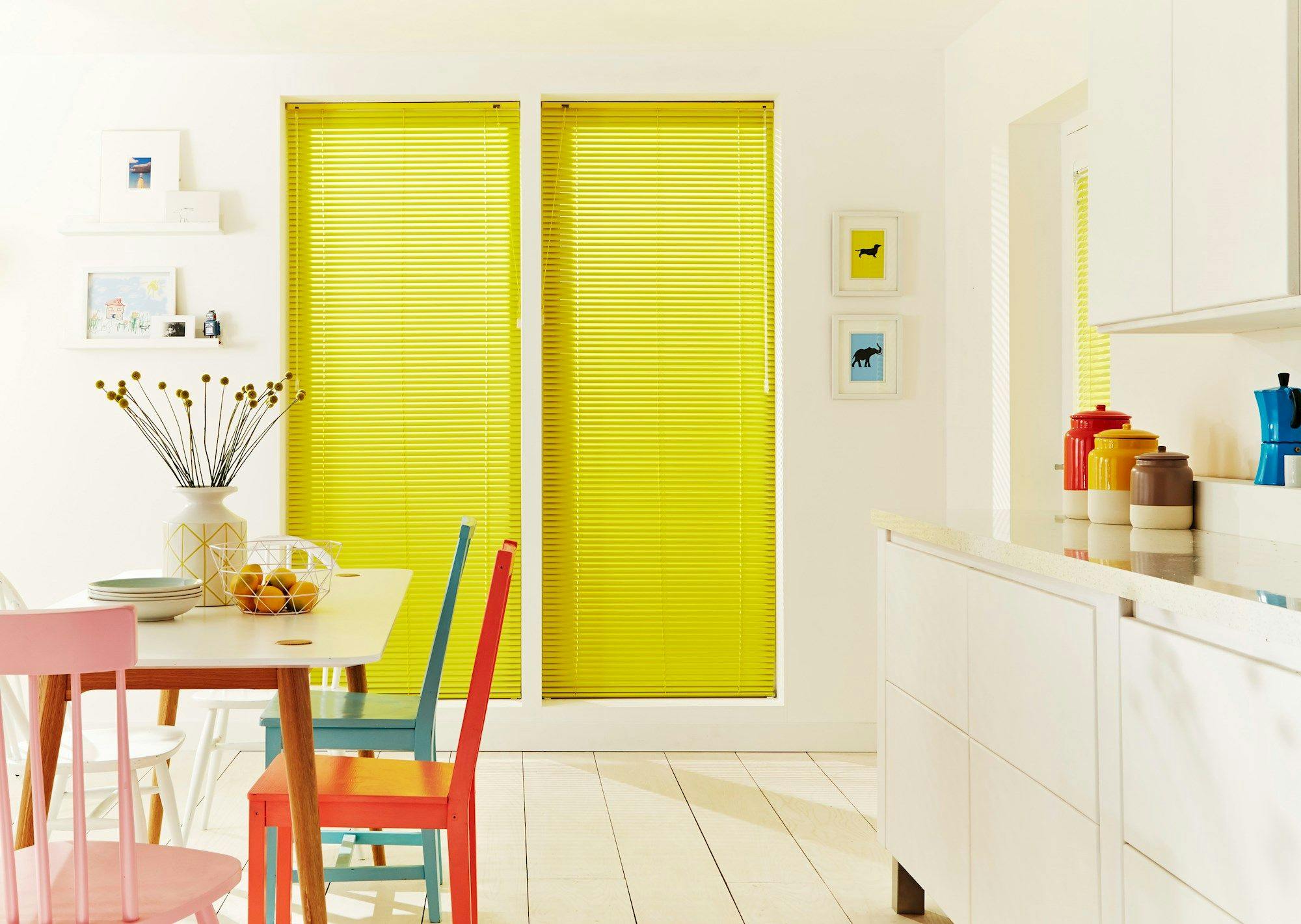 Yellow blinds cover large windows. A dining table with colorful chairs sits nearby, adorned with a vase of flowers and fruit bowl. Kitchen counter holds vibrant containers. White walls display framed art.