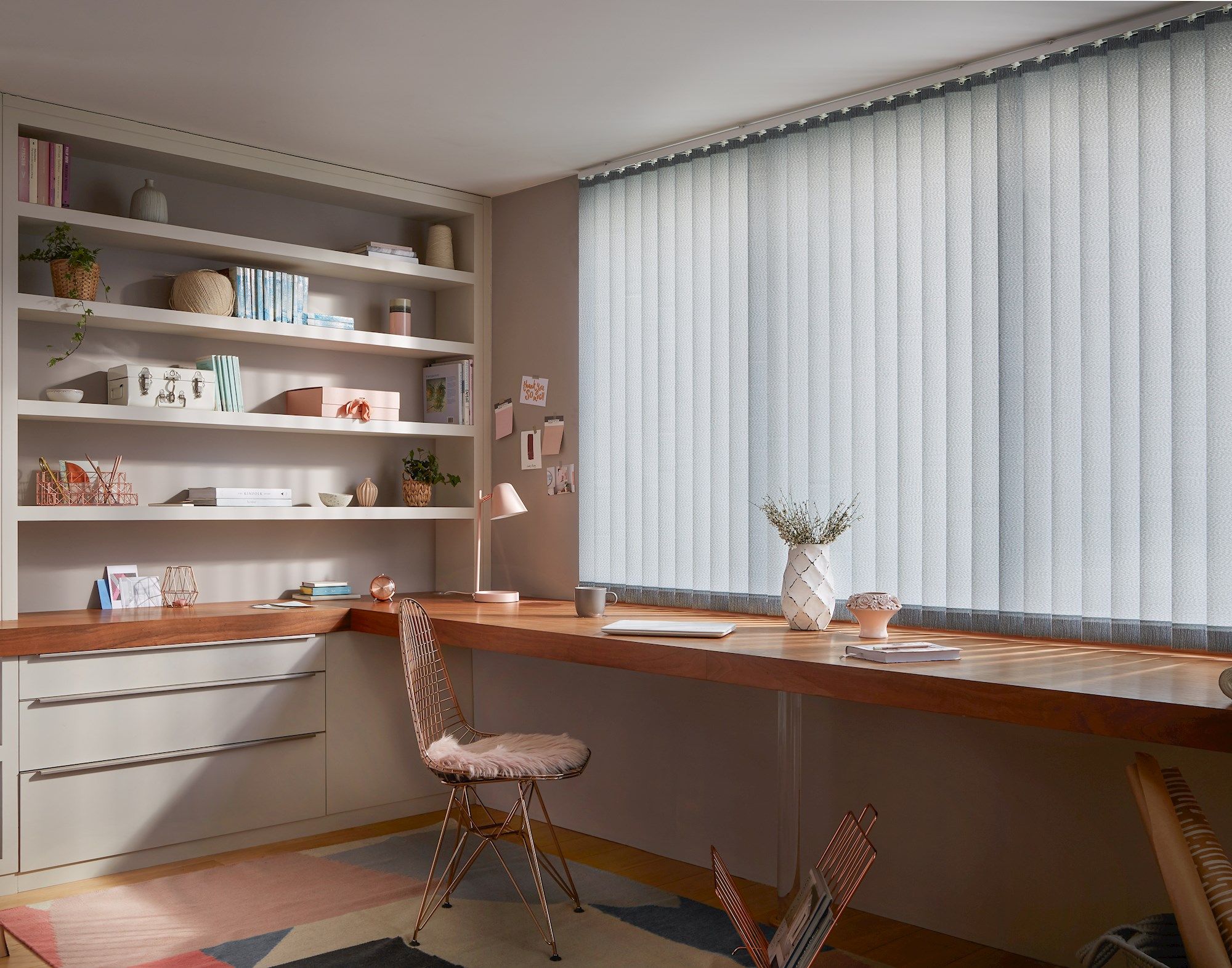 A wooden desk adorned with books, a lamp, and plants sits under vertical blinds. Shelves with decor and books fill the adjacent wall, creating a tidy, well-lit workspace.