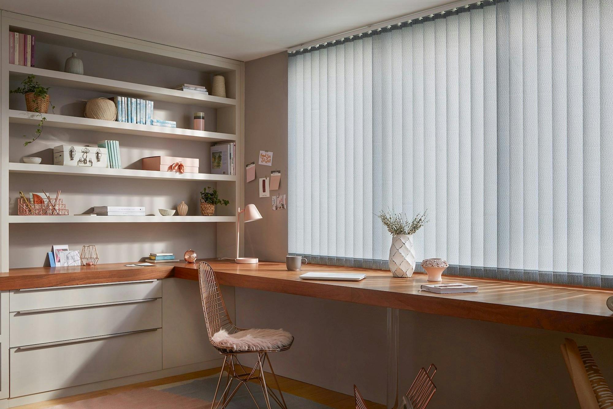 A wooden desk adorned with books, a lamp, and plants sits under vertical blinds. Shelves with decor and books fill the adjacent wall, creating a tidy, well-lit workspace.