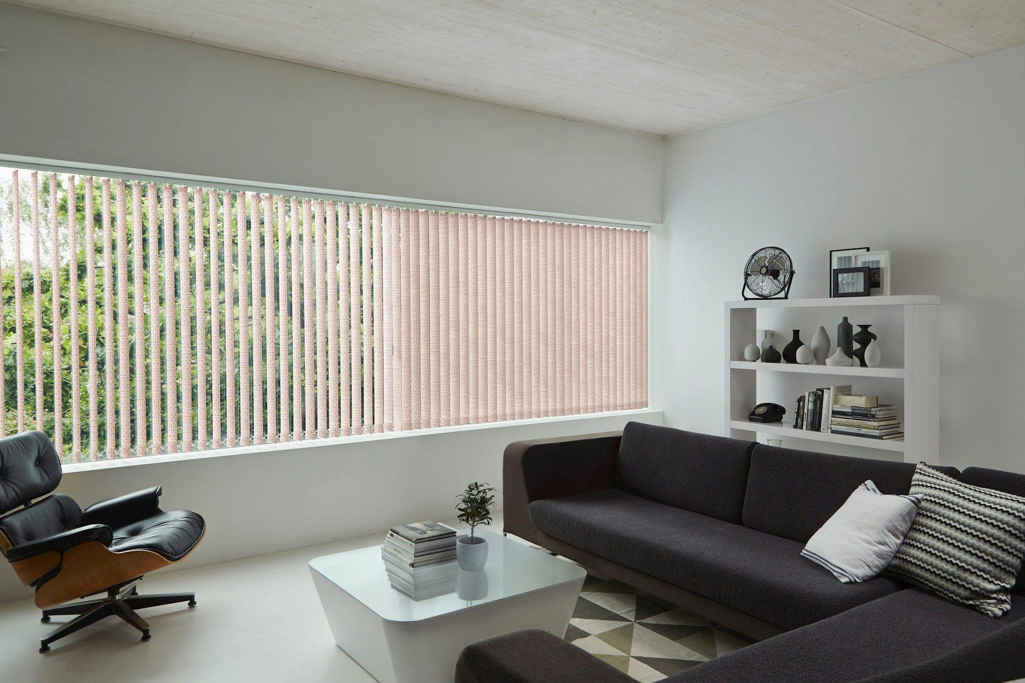 A black sectional sofa sits in a modern, minimalist living room. A coffee table with books and a small plant is in front, next to a bookshelf and an armchair by large vertical blinds.
