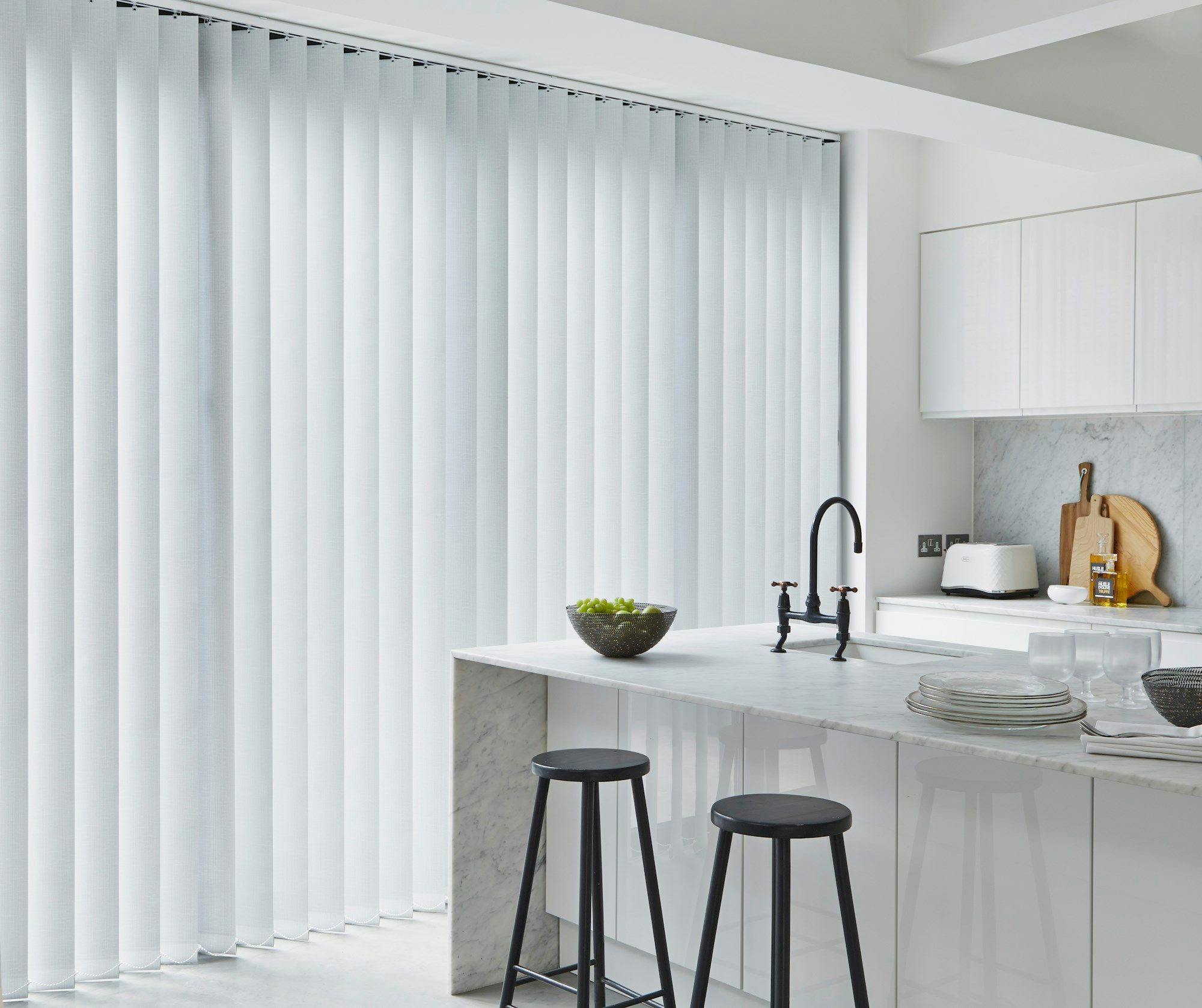 Vertical blinds cover a large window, partially open, in a modern kitchen with a marble countertop. Two black stools sit nearby, beneath a row of glossy white cabinets.
