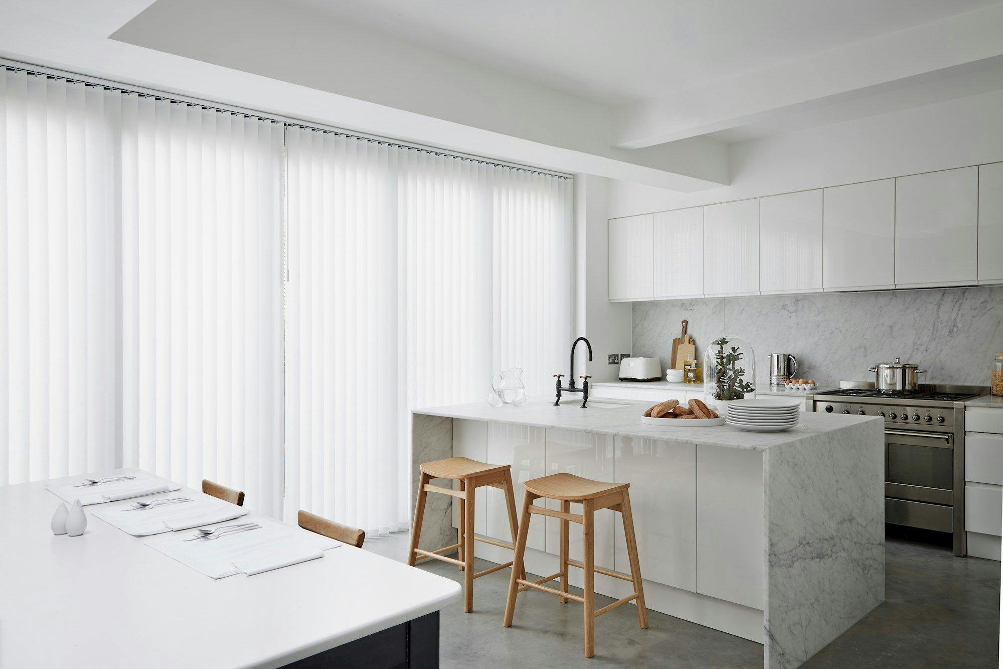 A modern kitchen features a marble island with two wooden stools, set in a bright room with vertical blinds, white cabinetry, and stainless steel appliances, including a stove and sink.