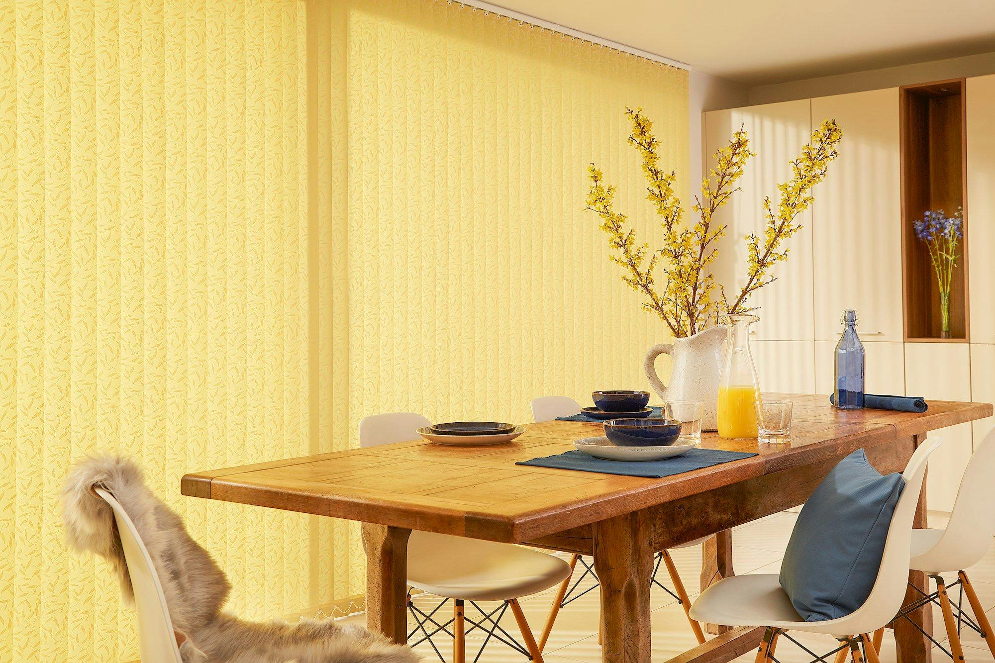 A wooden dining table set with blue bowls, pale yellow flowers in a pitcher, and a glass of orange juice, situated in a brightly lit room with yellow curtains.