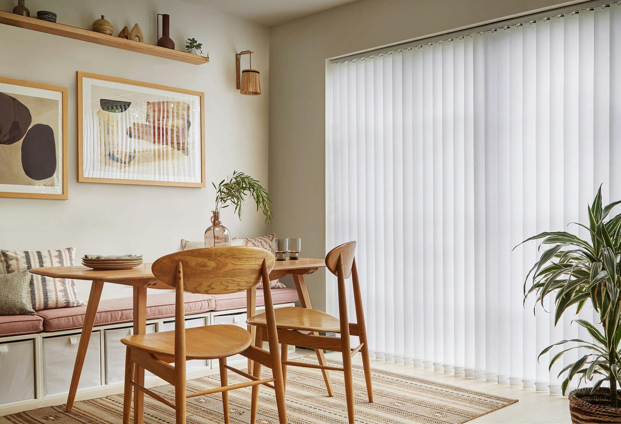 A wooden dining table with chairs is set in a cozy room, featuring wall art, cushions, and a large window with vertical blinds. Plants add a touch of greenery.