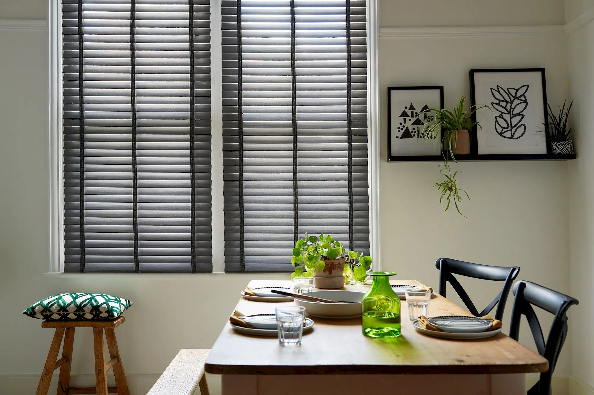 A sunlit dining room features a wooden table set with plates, glasses, and a green jug. Black chairs and a stool surround it, with blinds covering the windows and framed artwork and plants on shelves.