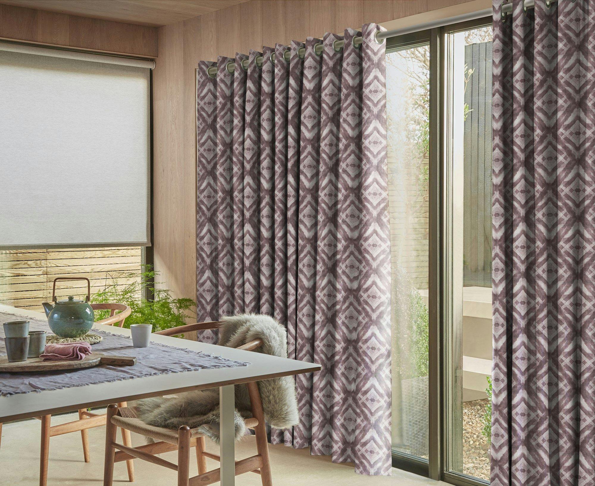 Curtains with geometric patterns hang in front of a glass door, allowing light into a modern room with a dining table set with dishes and a teapot.