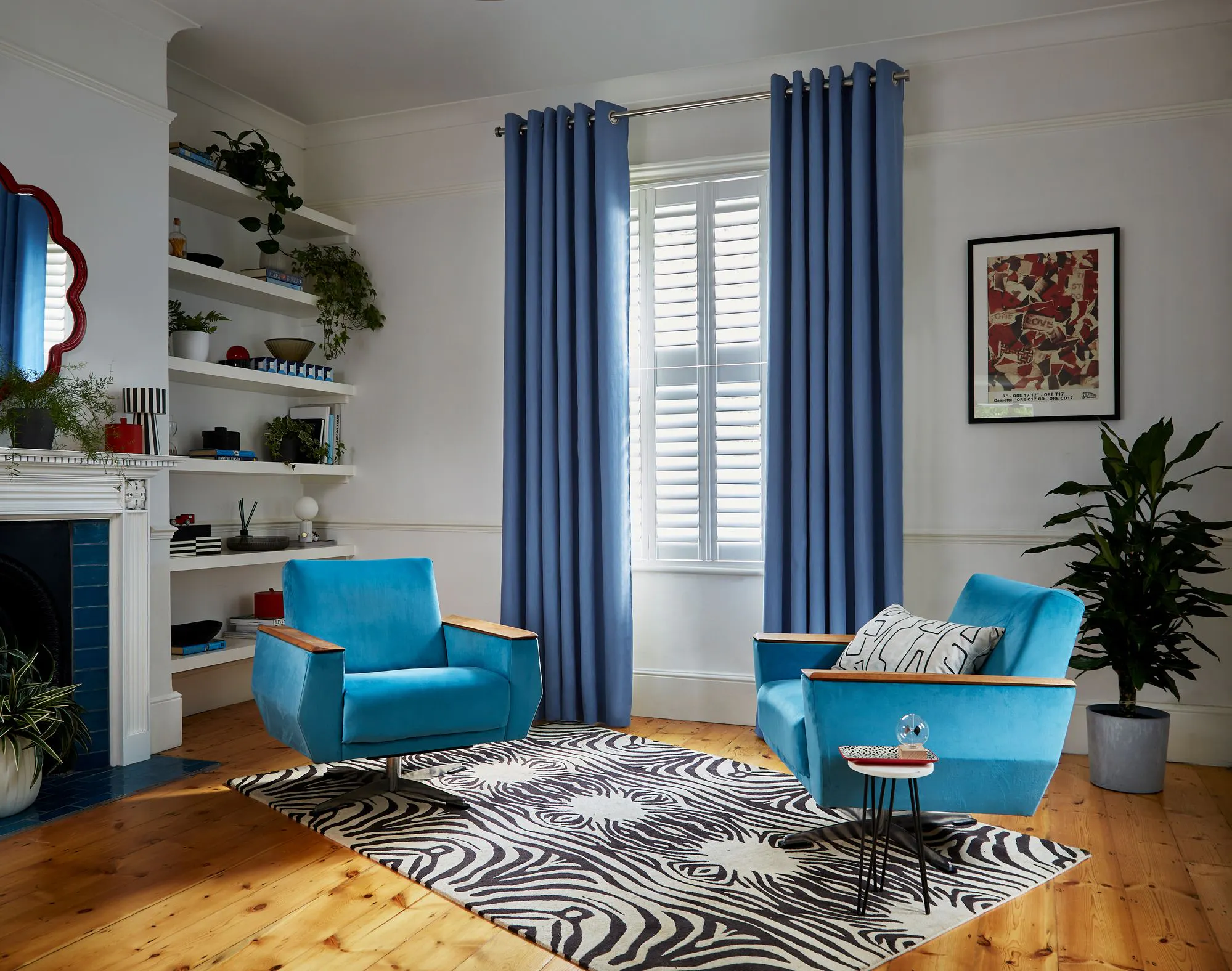 Two blue armchairs with wooden armrests sit on a zebra-patterned rug in a living room. Blue curtains cover the window, with plants and shelves on one side.