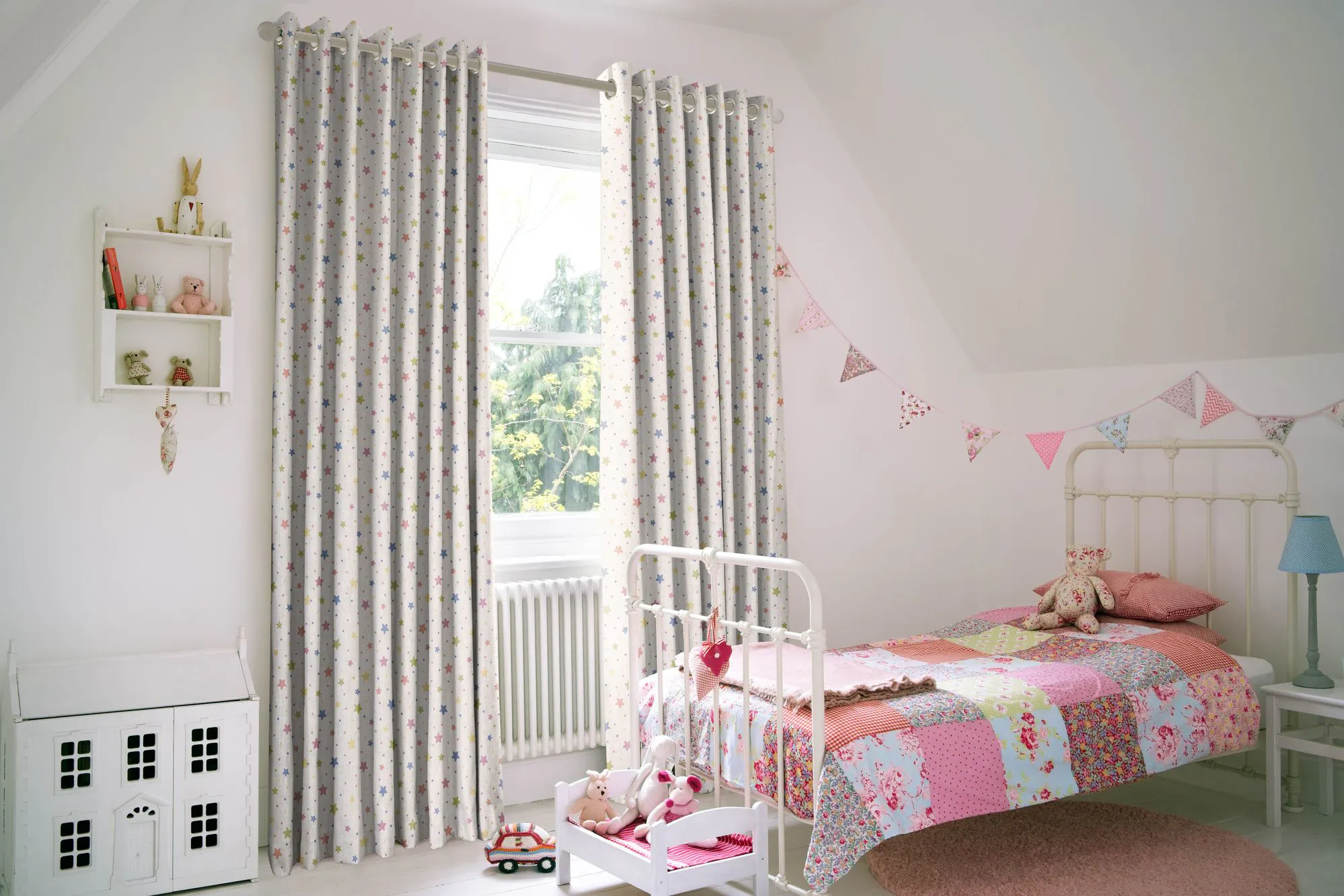 A child's bedroom features a white metal bed with a colorful patchwork quilt, toys, and bunting on the wall. Curtains with patterns frame a window overlooking greenery.