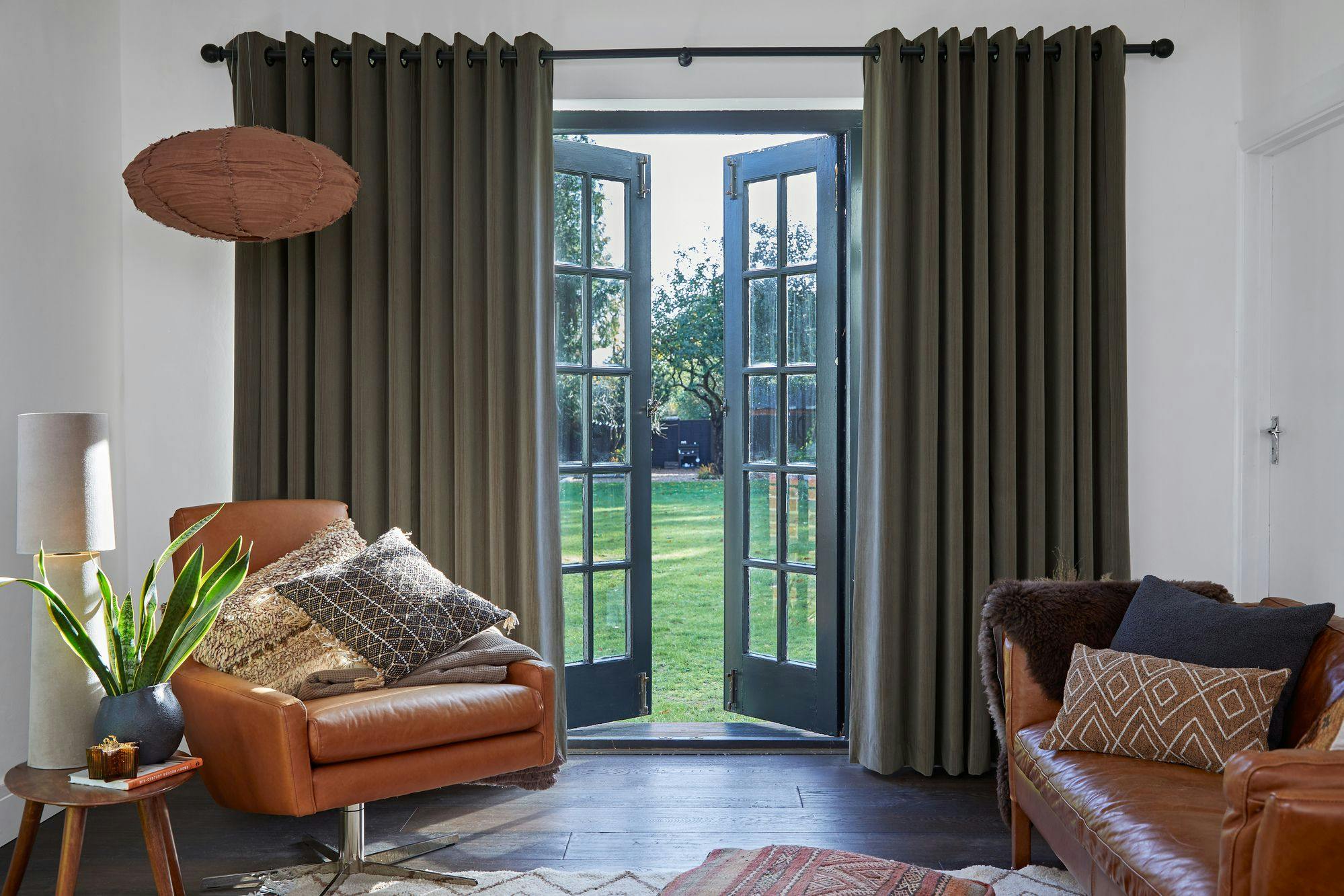 Armchair holds textured pillows, beside a side table with a plant and lamp. French doors open to a garden, surrounded by dark curtains, creating a cozy, inviting setting.