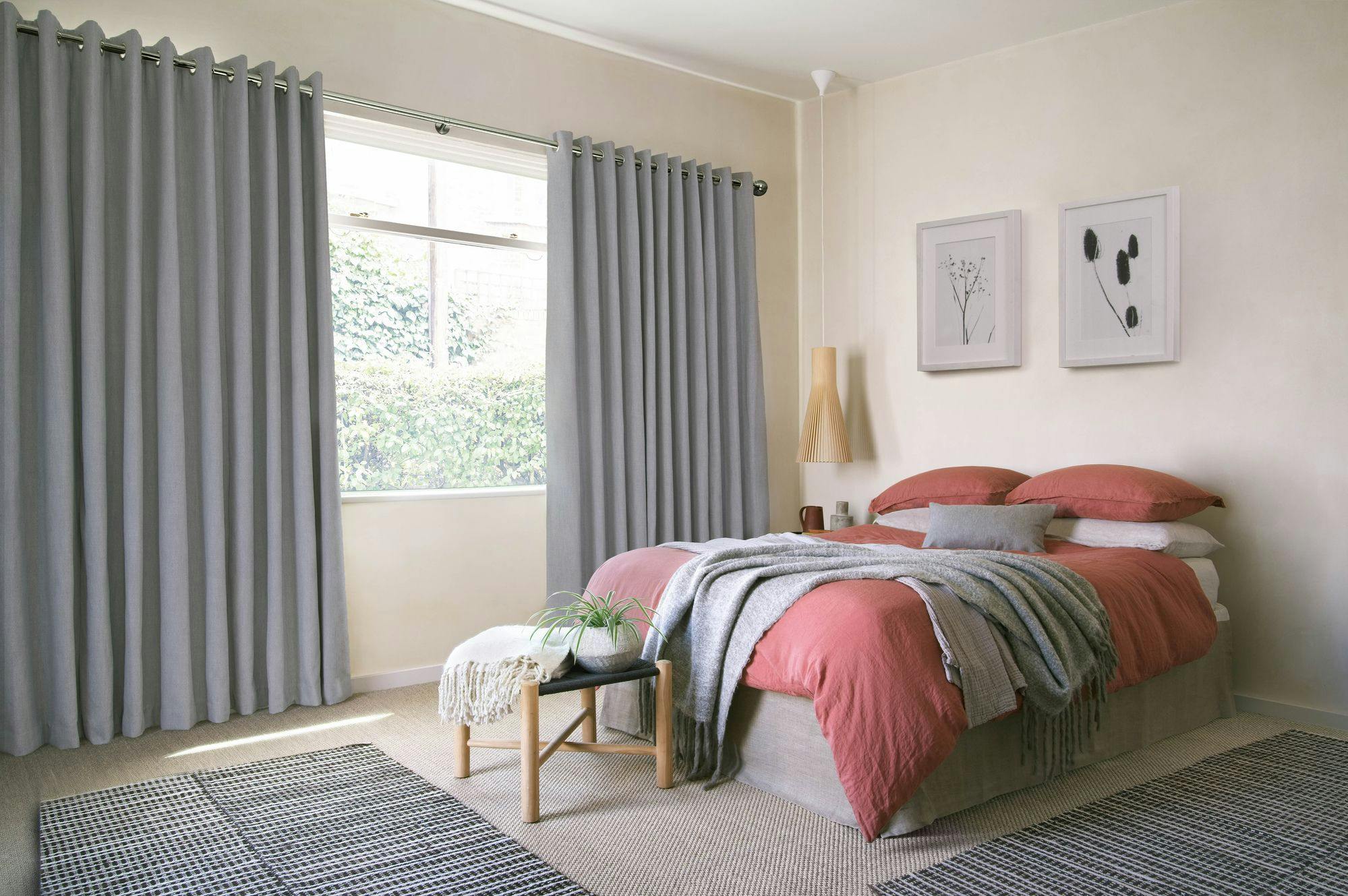 Bed with coral bedding sits in a cozy bedroom, surrounded by gray curtains and light walls. A small bench holds a plant and blanket, enhancing the inviting atmosphere.
